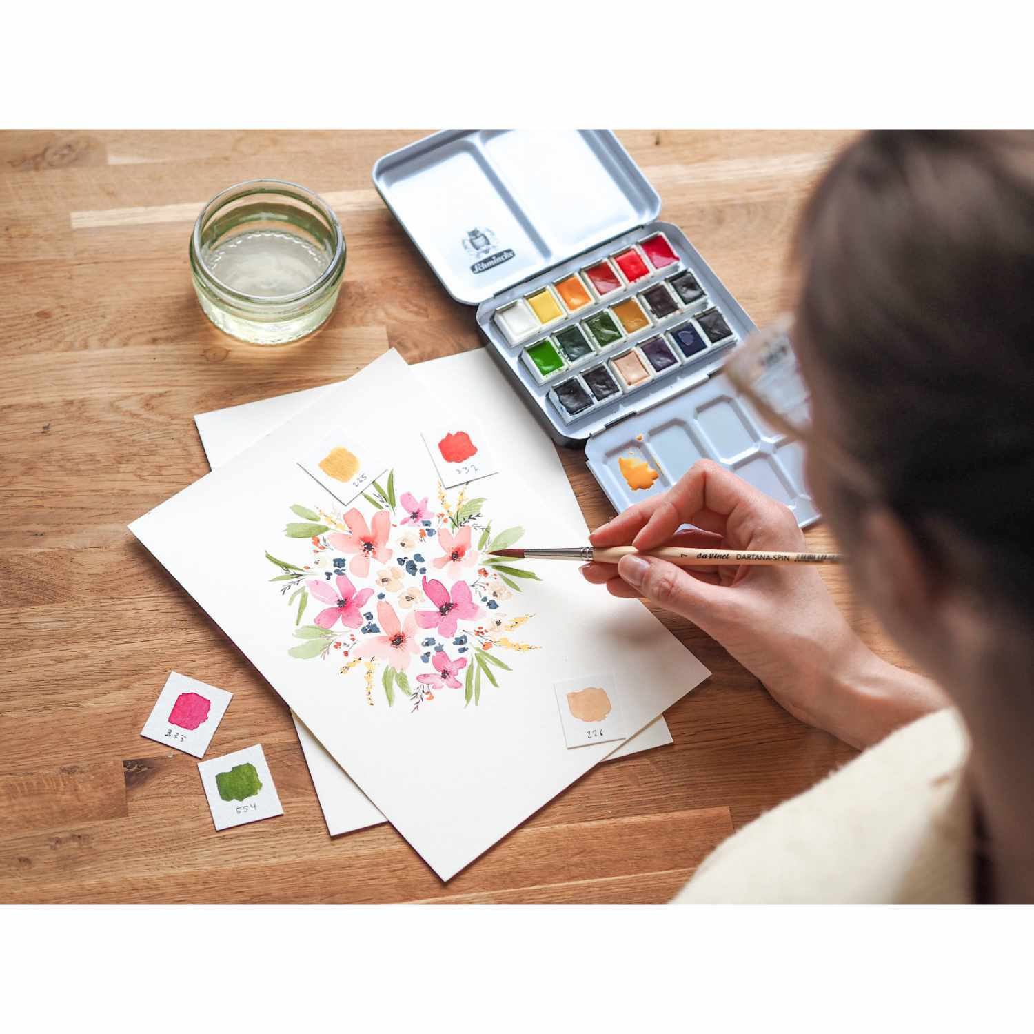 Schmincke Akademie Aquarellkasten mit 18 Farben, ideal für kreative Blumenmotive. Künstler malt mit Pinsel auf Papier, umgeben von Farbkarten.