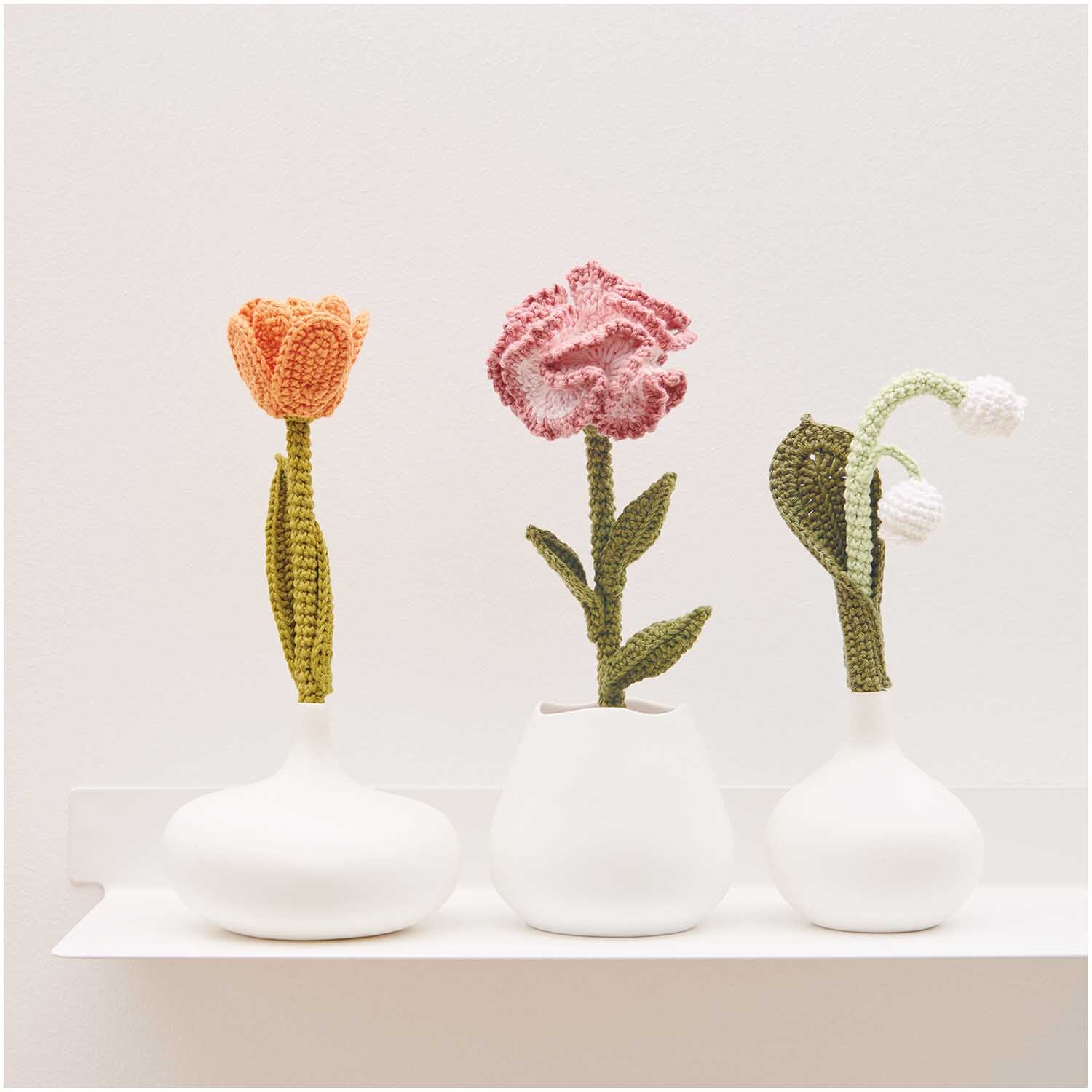 Creative Ricorumi dk: Drei gehäkelte Blumen in weißen Vasen auf einem Regal, dekorativ arrangiert.