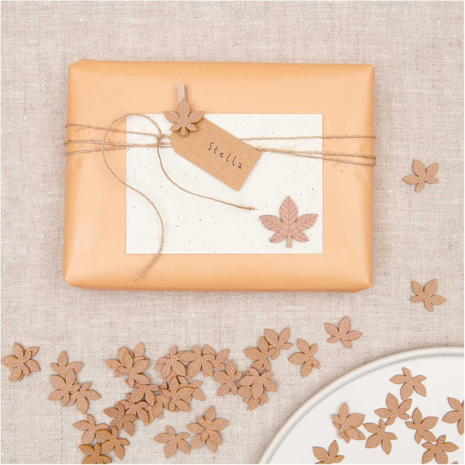 Holzsticker Blatt braun 12 Stück dekorieren ein Geschenk mit Etikett auf beigem Stoff. Ideal für kreative Bastelprojekte.