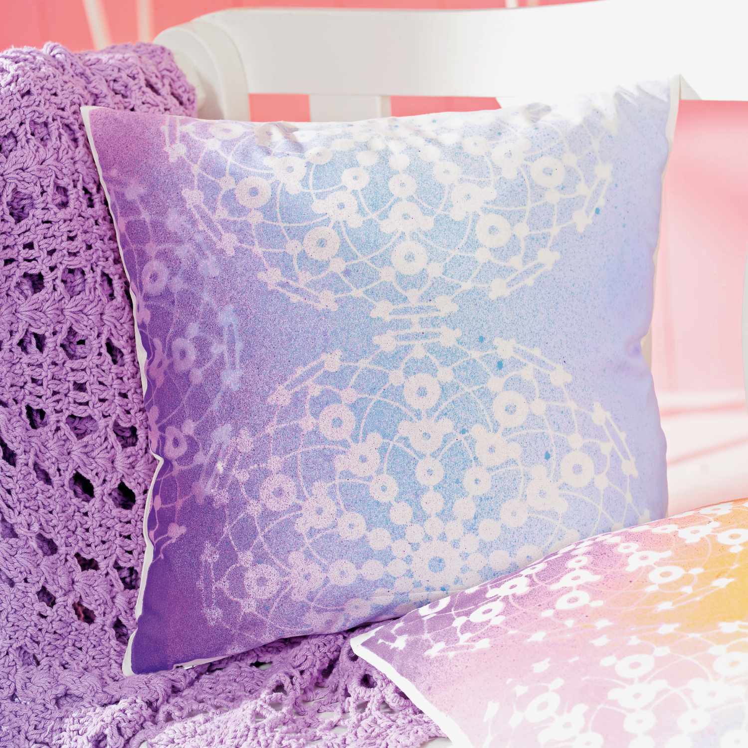 Fashion Spray: Dekoratives Kissen mit floralem Muster in Pastelltönen auf lila Häkeldecke.
