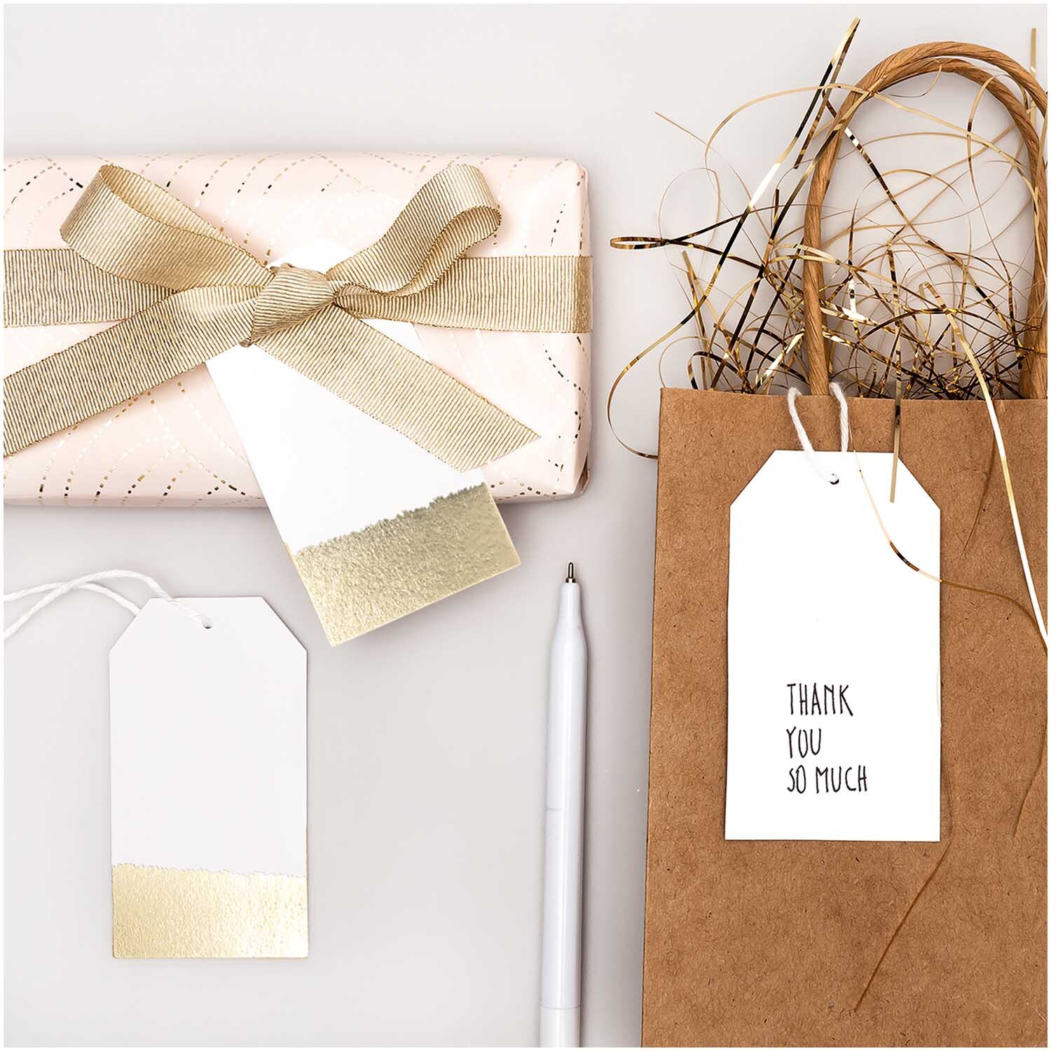 Paper Poetry Papieranhänger 4,5x8cm 24 Stück in Weiß-Gold neben Geschenk und Tüte. Ideal für kreative Verpackungen.