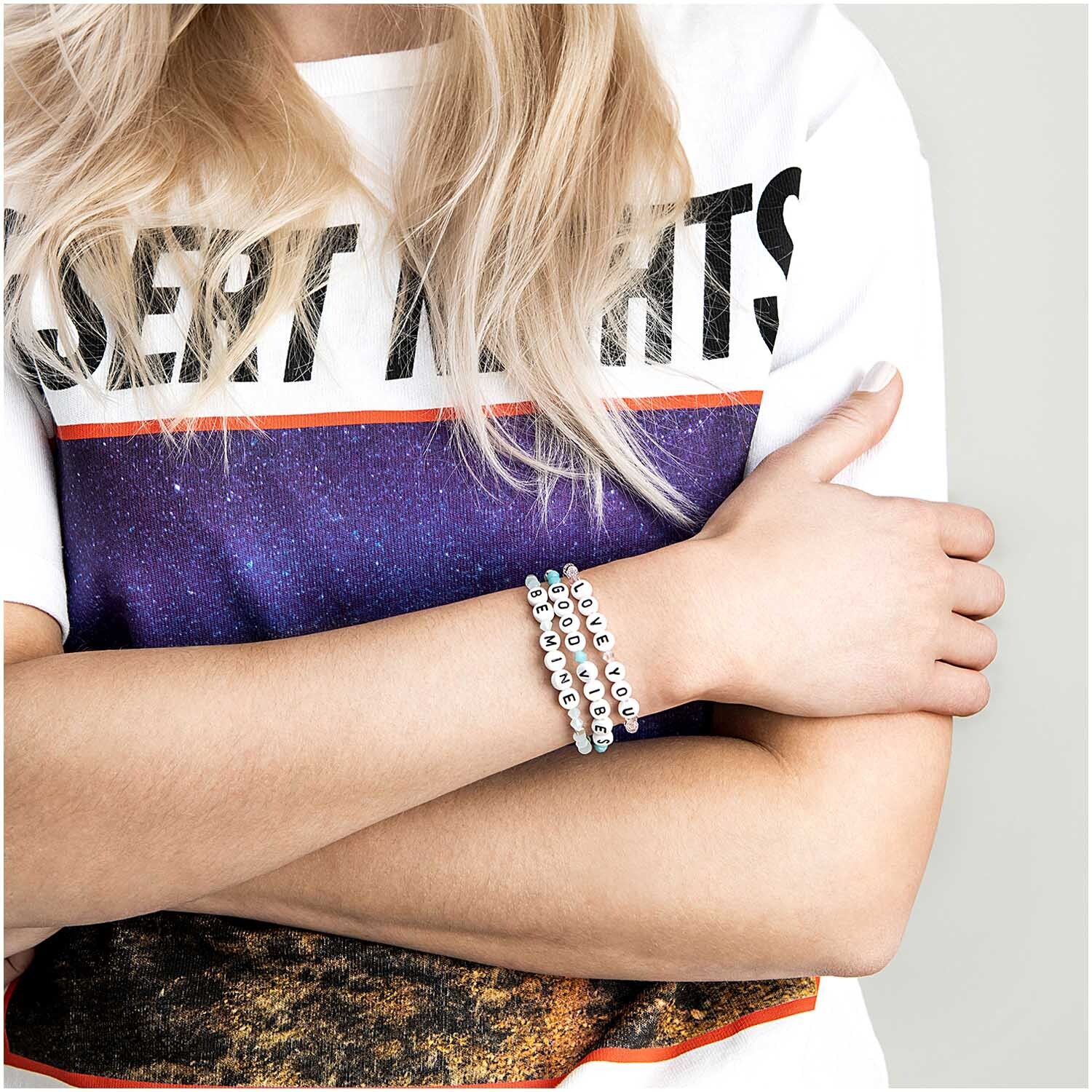 Buchstabenperle: Frau mit weißem T-Shirt und Armbändern von Rico Design, die Arme verschränkt.