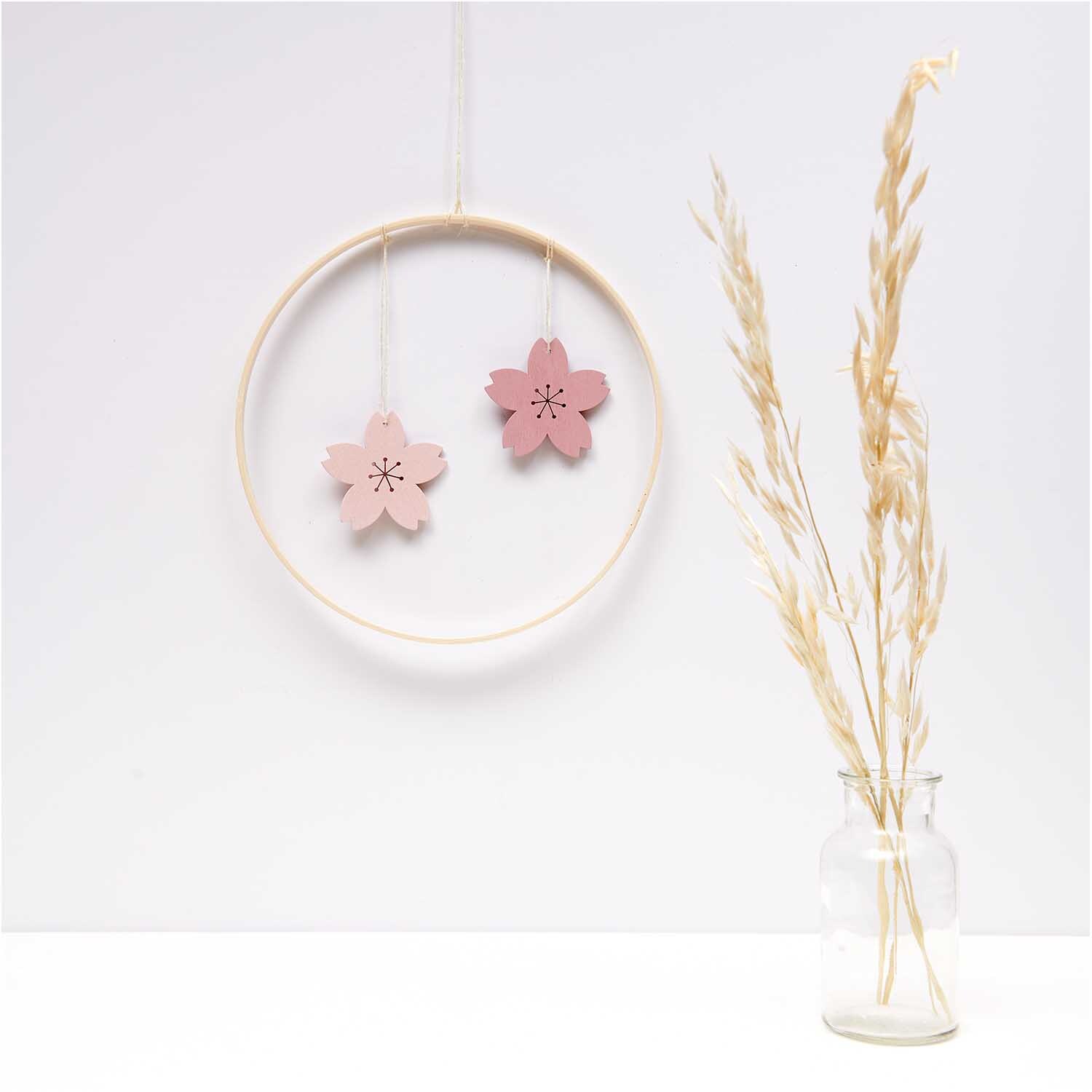 Hänger Blüte puder 1 Stück: Zwei rosa Blütenanhänger von Rico Design in einem Holzring, neben einer Vase mit Trockenblumen.