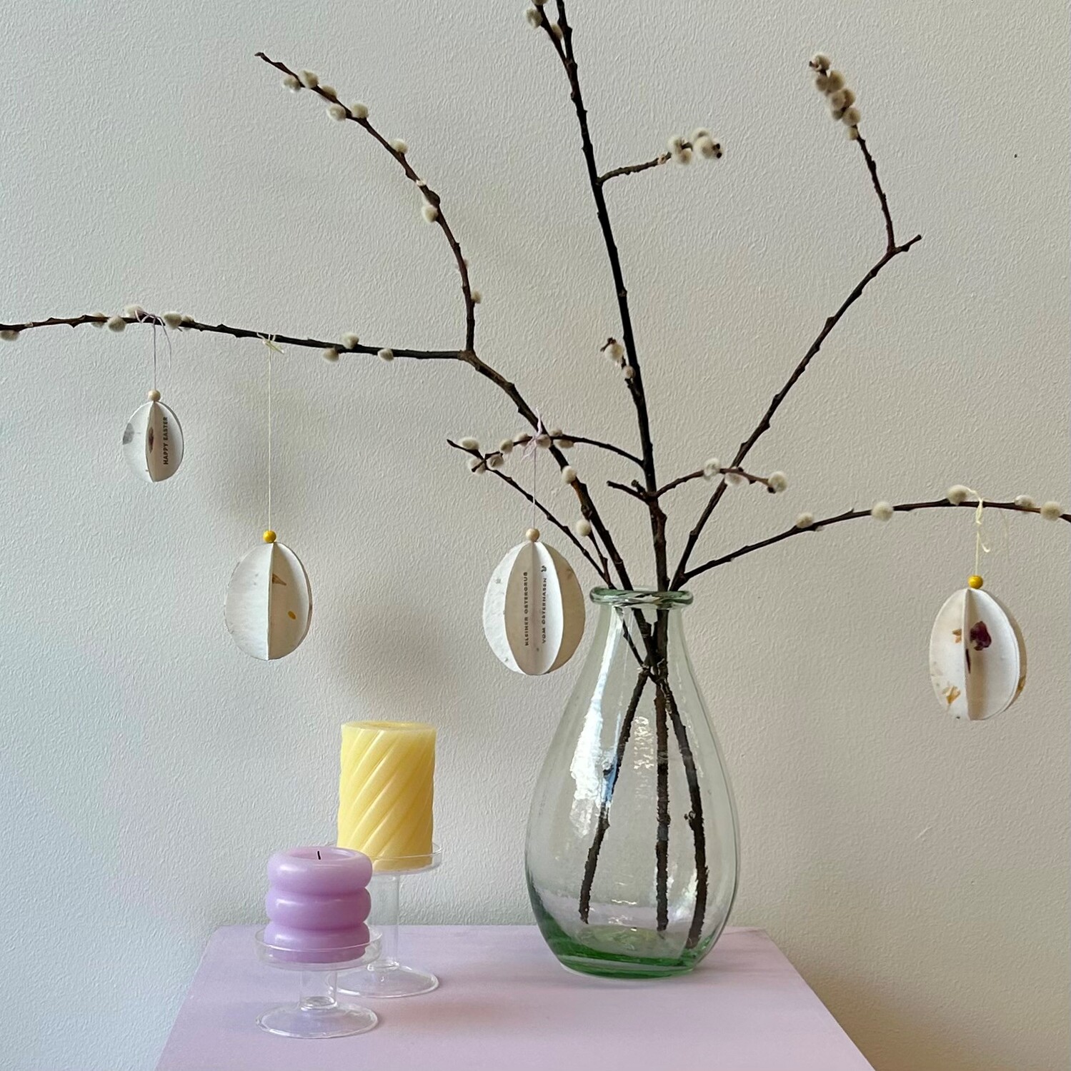 Bastelanleitung Osterei aus Papier: Zweige in Vase mit hängenden Papier-Ostereiern, daneben Kerzen.