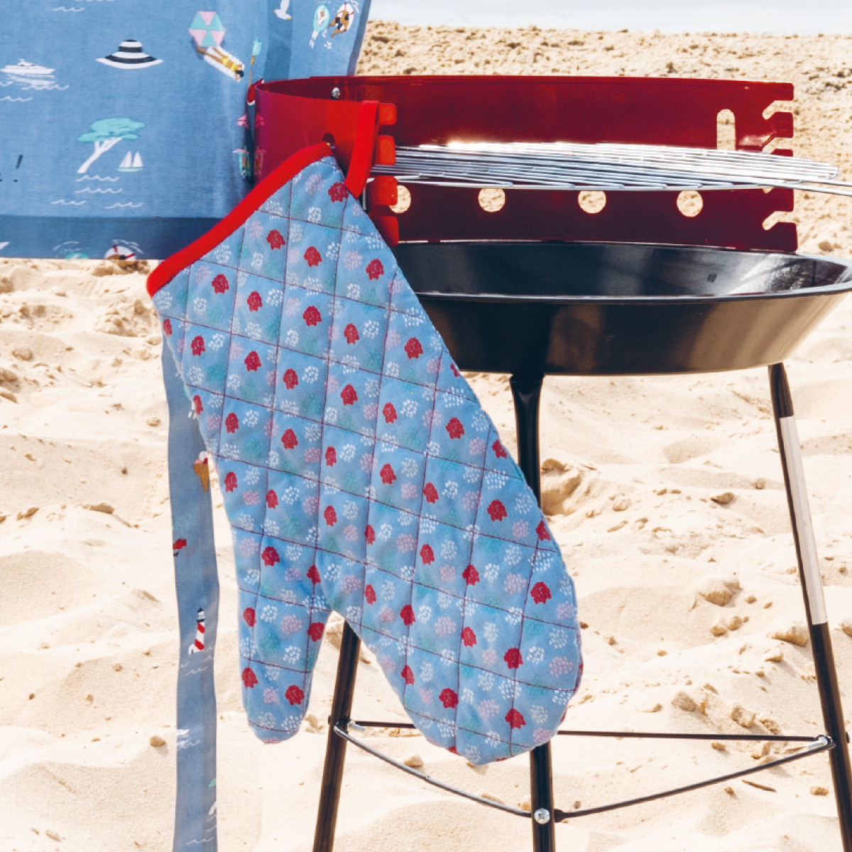 Nähanleitung Grillhandschuh: Ein blauer Grillhandschuh mit rotem Rand hängt an einem Grill am Strand.