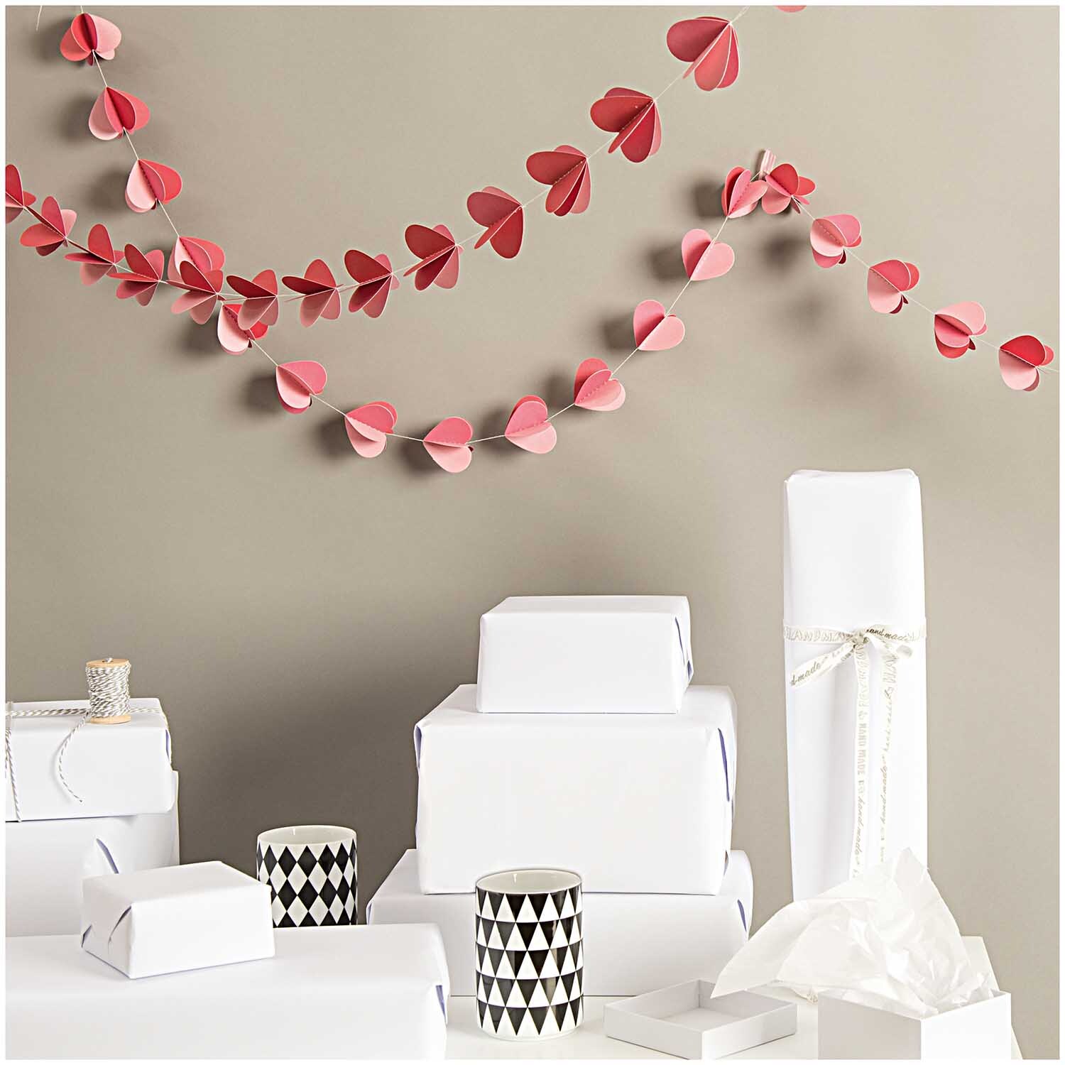 Girlande Herzen rot 1,5m von Rico Design schmückt eine Wand über weißen Geschenken und Kerzen.