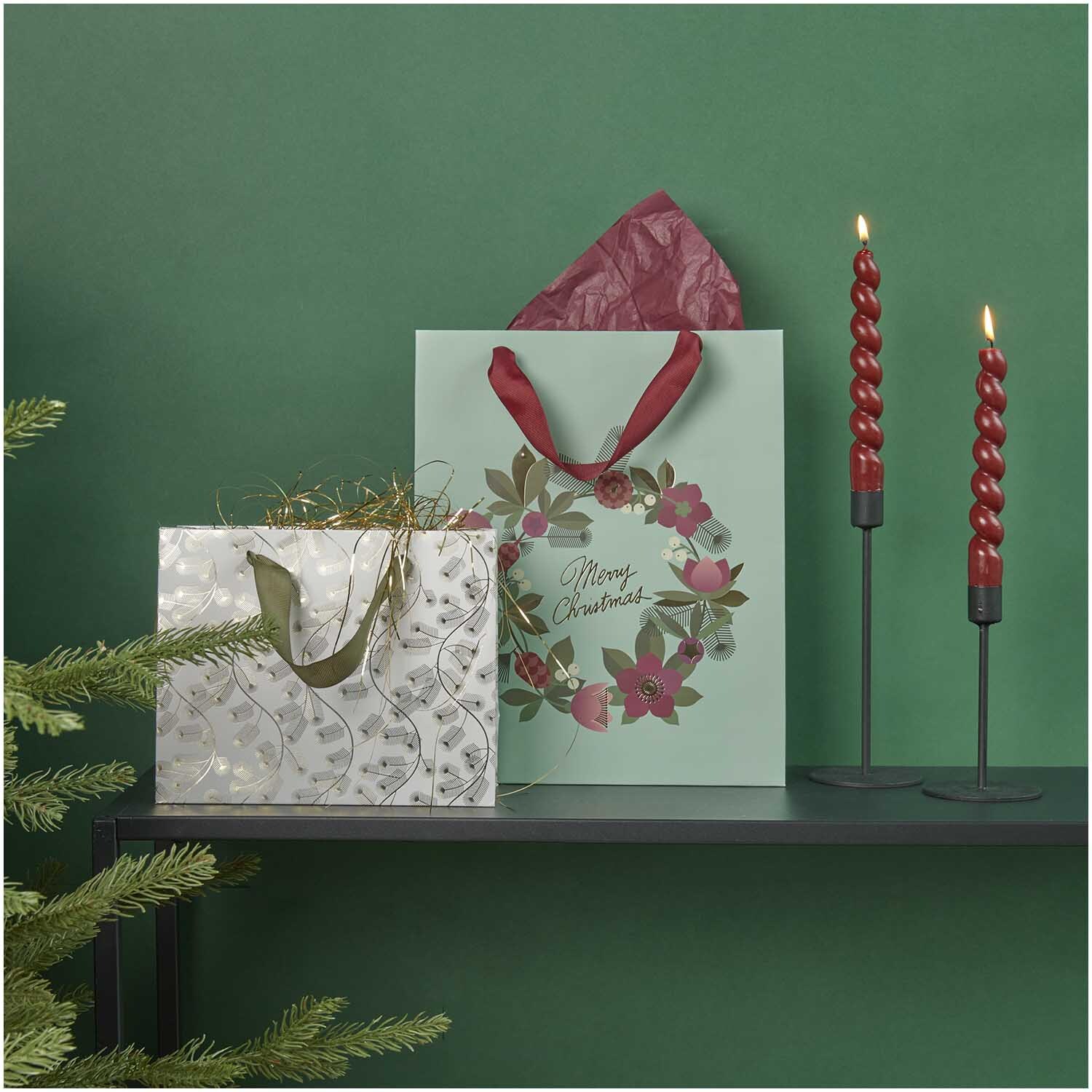 Paper Poetry Geschenktüte mit Kranzmotiv, dekoriert mit roten Kerzen und Tannenzweigen auf grünem Hintergrund.