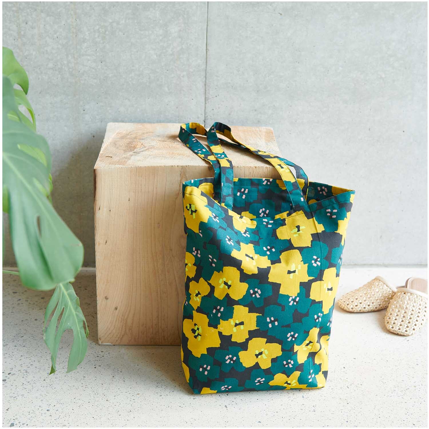Shopper Okina Hana Blumenprint 38x40x13cm, gelb-blaues Design, steht neben Holzwürfel und Pflanzenblatt. Ideal für stilvolle Einkäufe.