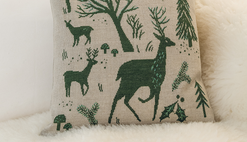 Stickanleitung Weihnachtliches Kissen gestalten: Kissen mit grünen Waldmotiven und Rehen auf beigem Stoff, ideal für festliche Dekoration.