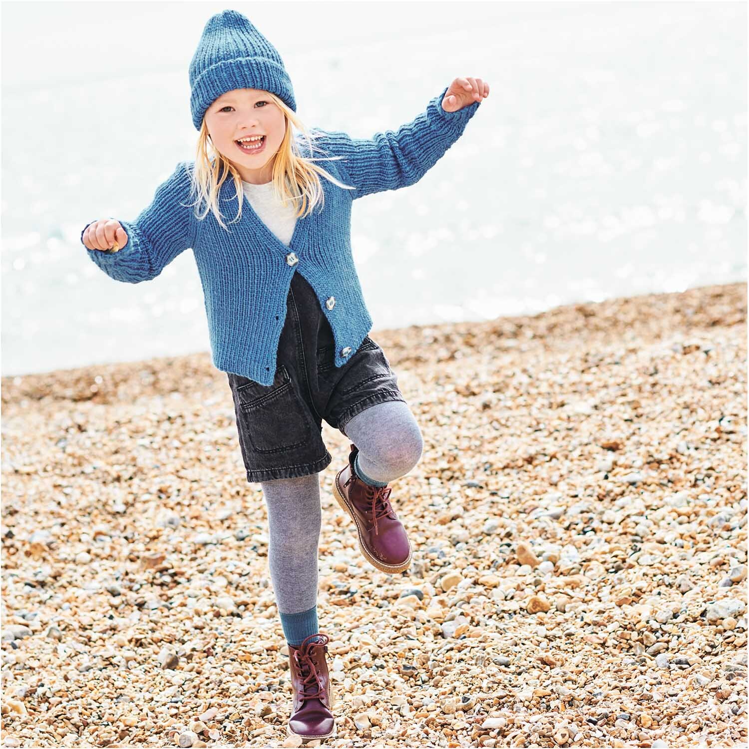 Essentials Super aran: Kind in gestrickter blauer Kleidung am Strand, trägt Mütze und Strickjacke.