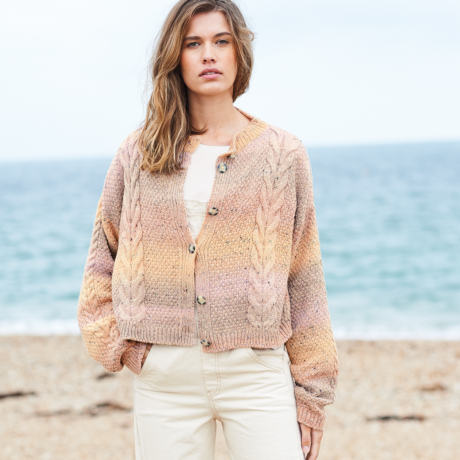 Strickanleitung Tweedjacke aus Fashion Cotton Light & Long Tweed dk: Frau am Strand in pastellfarbener Strickjacke mit Zopfmuster, beige Hose.