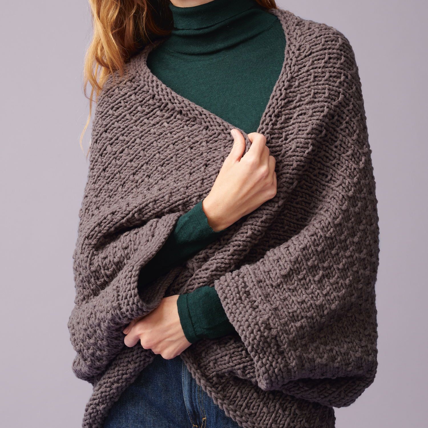 Strickanleitung Poncho aus Polar: Frau trägt einen grob gestrickten, grauen Poncho über einem grünen Rollkragenpullover.