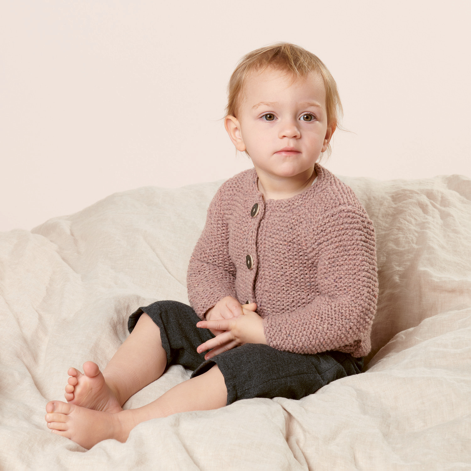 Strickset Babyjanker Modell 16 aus Trachtenbuch I mog di: Kleinkind in rosa Strickjacke auf beigem Stoff sitzend.