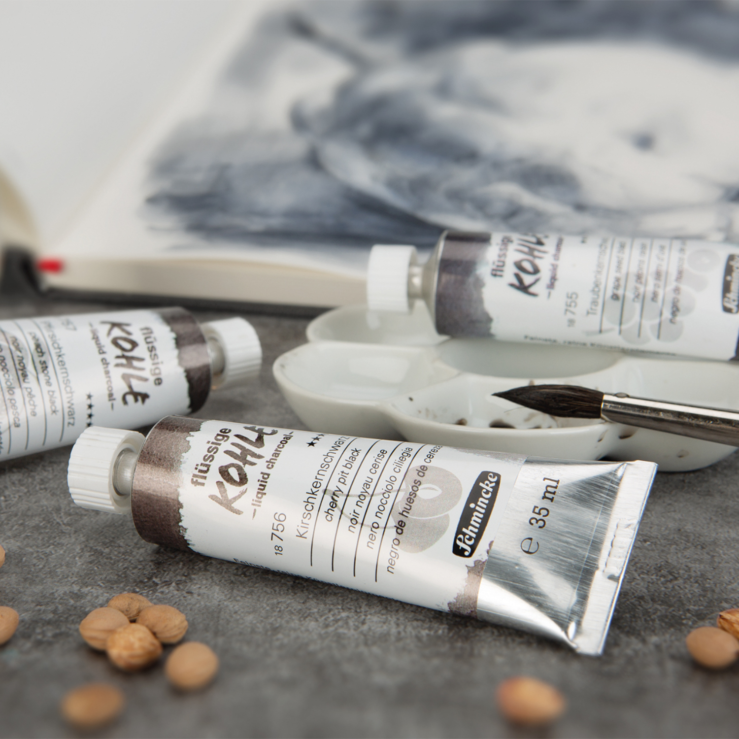 Pigmente Trio flüssige Kohle Set, Schmincke: Drei Tuben mit 3x5ml Farben auf grauem Tisch, Pinsel und Palette im Hintergrund.