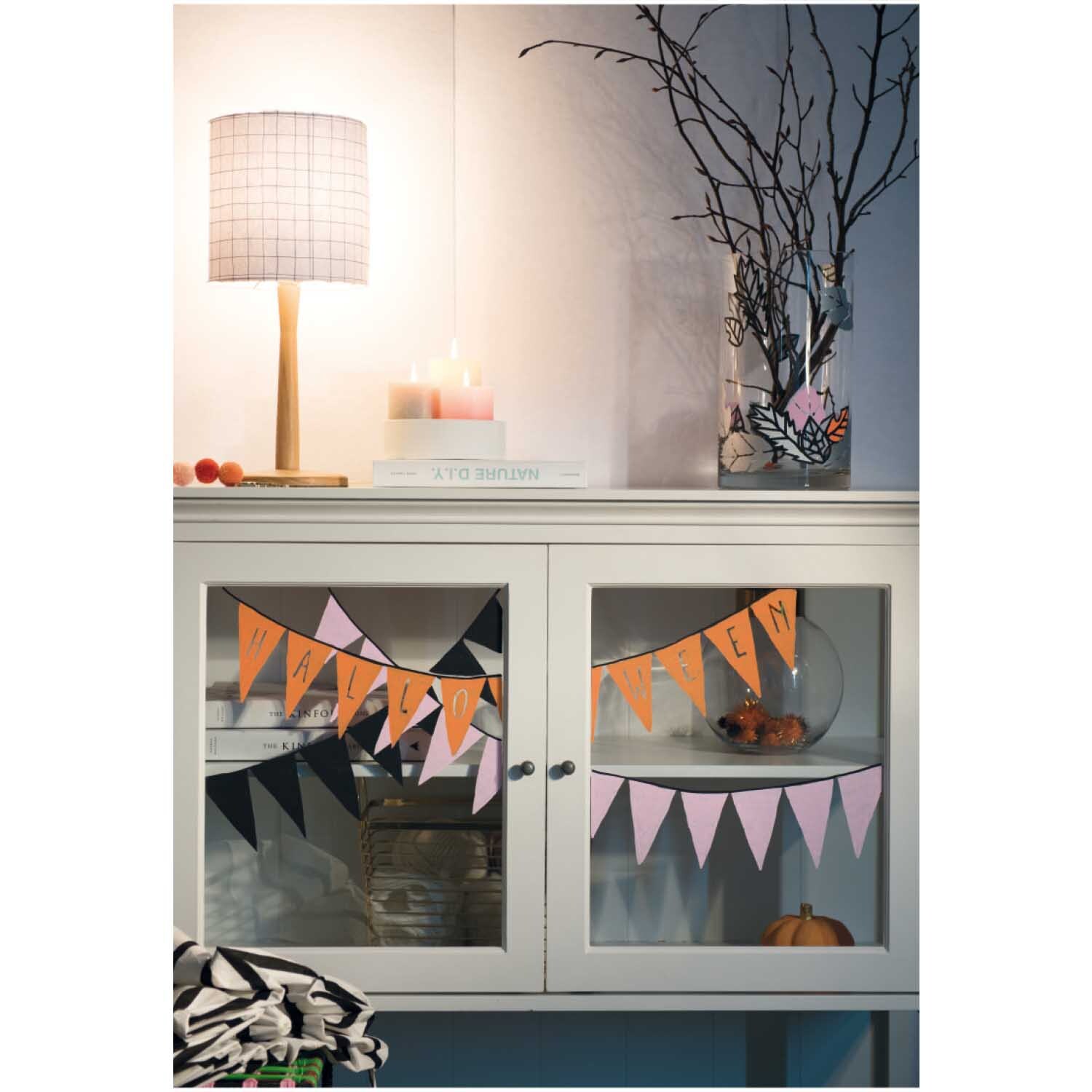 Vorlagenmappe Halloween Fenster- und Glasmalerei mit Kreidestiften: Bunte Girlanden schmücken ein weißes Sideboard, dekoriert mit Kürbissen und Zweigen.
