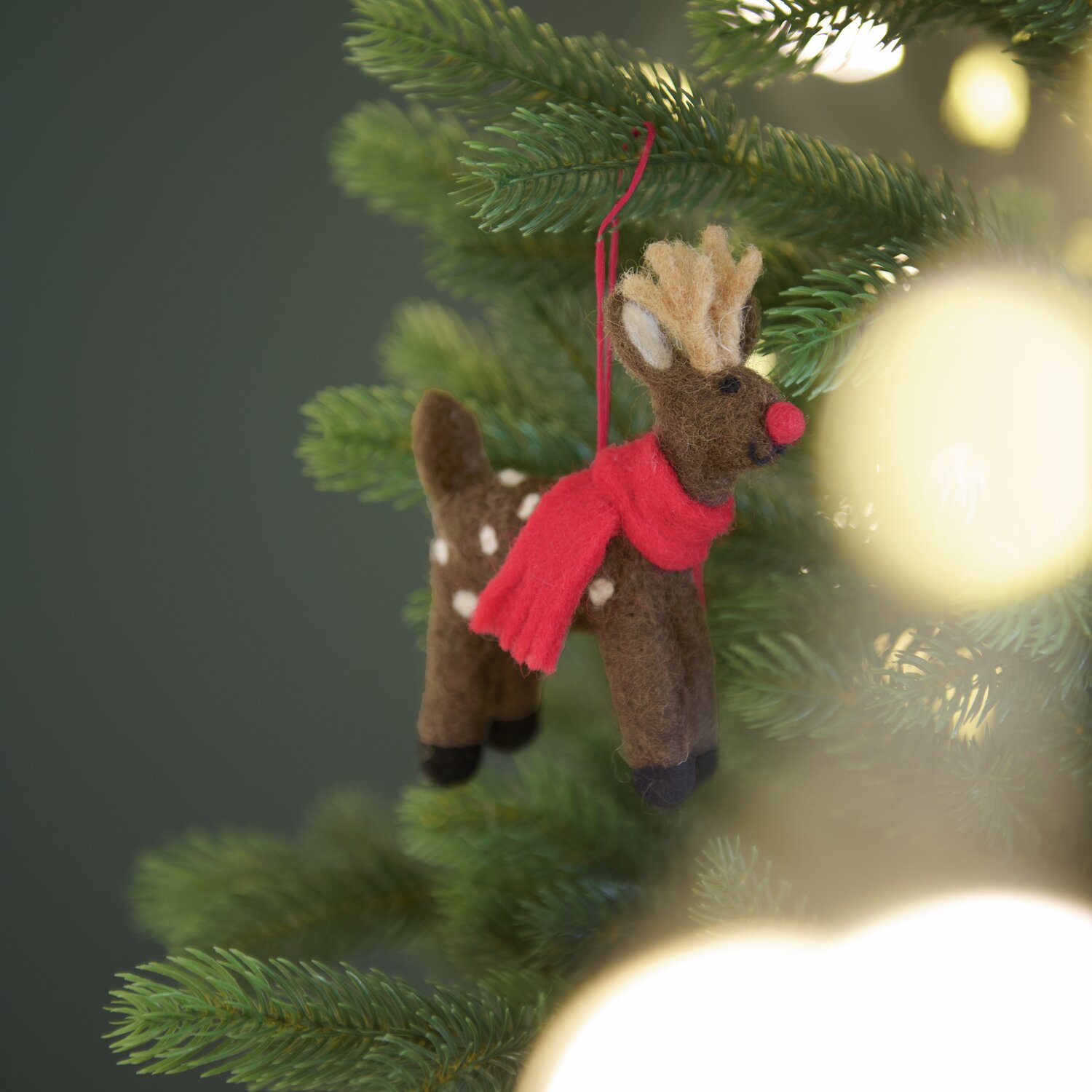 Aufhänger Filz-Hirsch mit Schal: Dunkelbrauner Hirsch aus Filz mit rotem Schal, hängend an einem Tannenbaum. Größe 12x8x2,5cm.