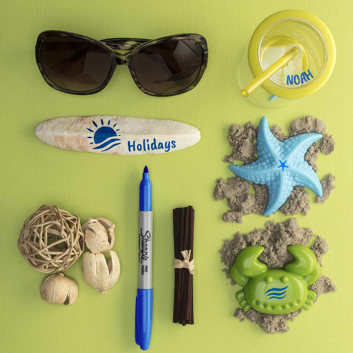 Permanentmarker 0,9mm von Sharpie in Blau neben Sonnenbrille, Muscheln und Stranddekoration auf grünem Hintergrund.