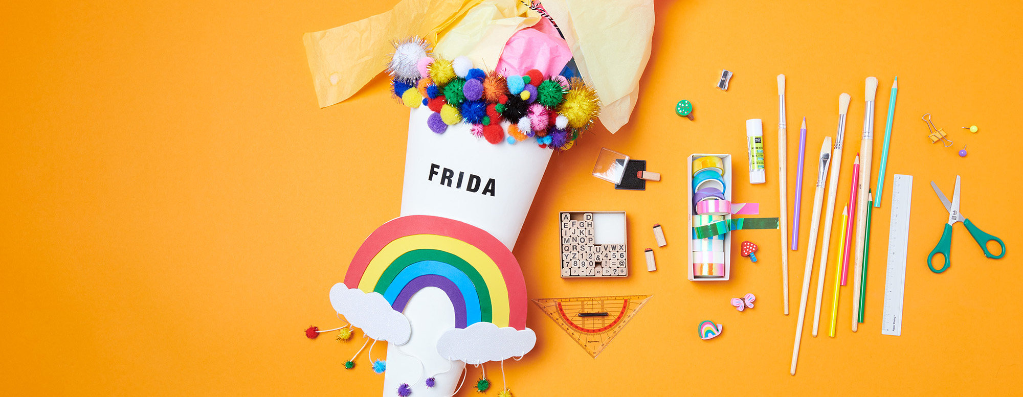 Weiße Schultüte mit Regenbogen und bunten Pompons, daneben liegen Stifte, Pinsel, Stempel, bunte Tapes, Radiergummis, ein Lineal und eine Schere. Flatlay von Produkten.