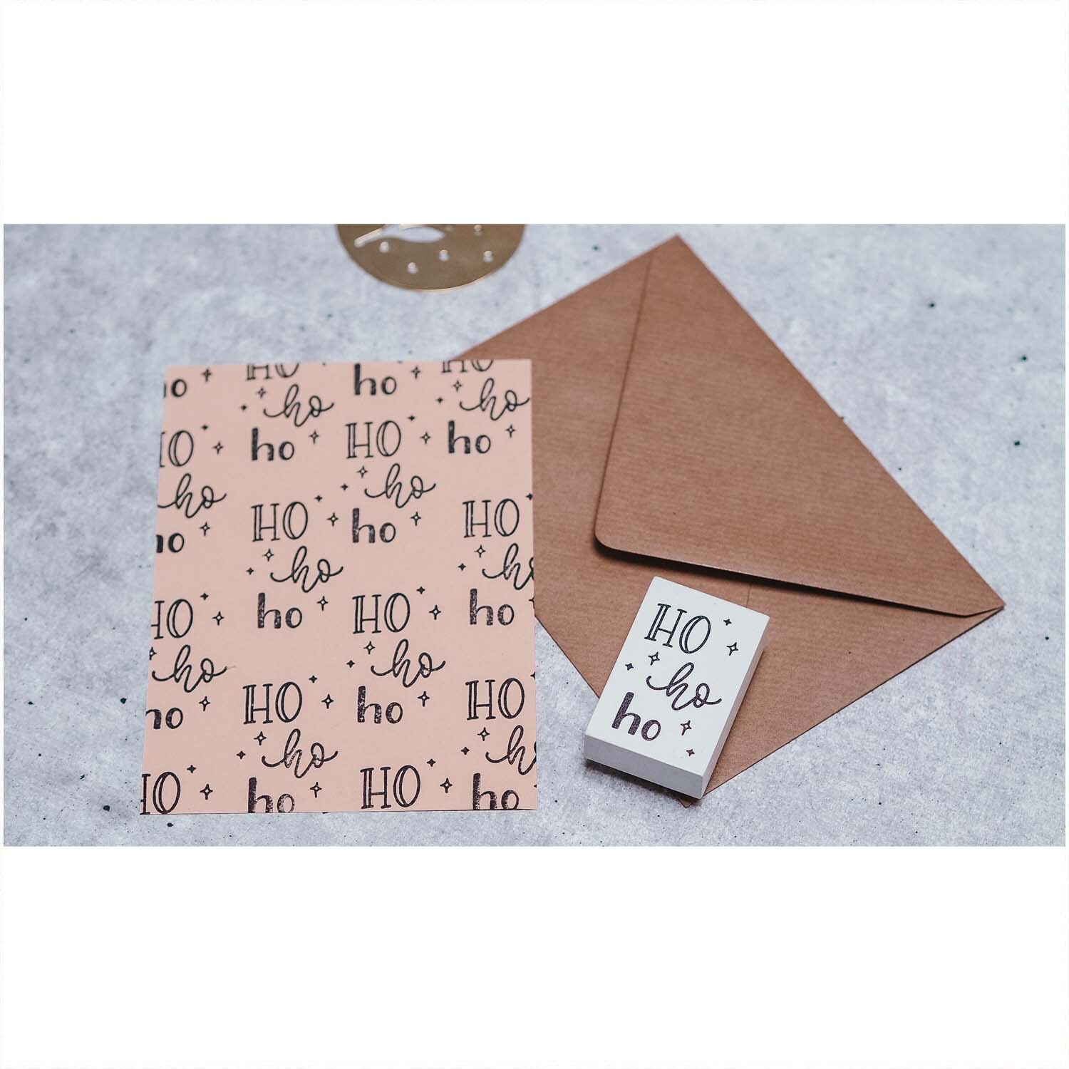 Stempel Ho ho ho weiß 35x55mm neben braunem Umschlag und Karte mit gleichem Design auf grauem Hintergrund.