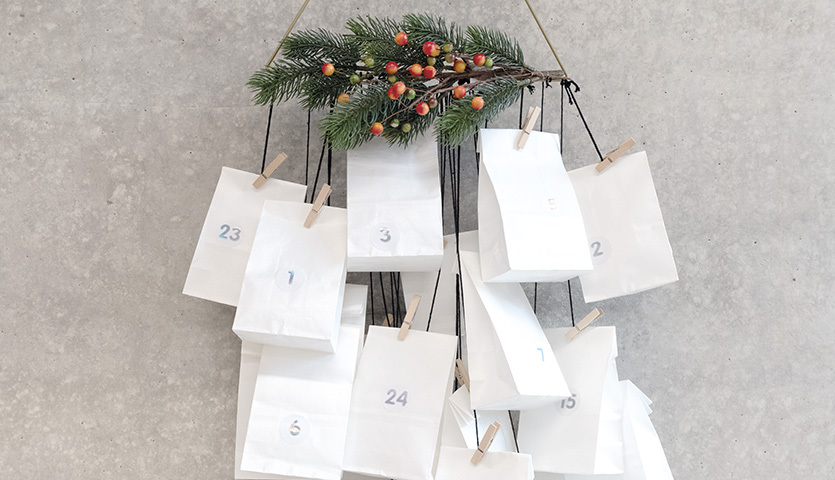 Bastelanleitung moderner Papiertüten-Adventskalender: Weiße Tüten mit Zahlen, dekoriert mit Tannenzweigen und roten Beeren, an einer Wand hängend.