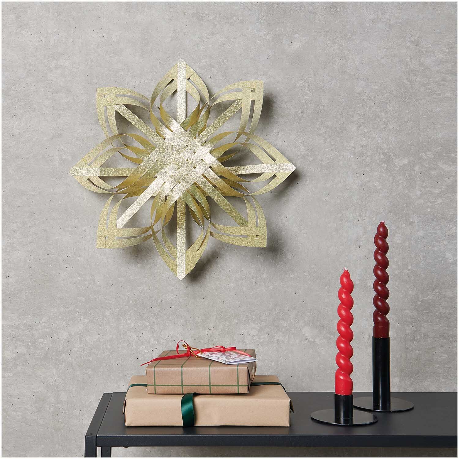 Paper Poetry Web-Stern Größe M 270g/m2 in funkelndem Gold an einer grauen Wand, dekoriert mit Geschenken und roten Kerzen.
