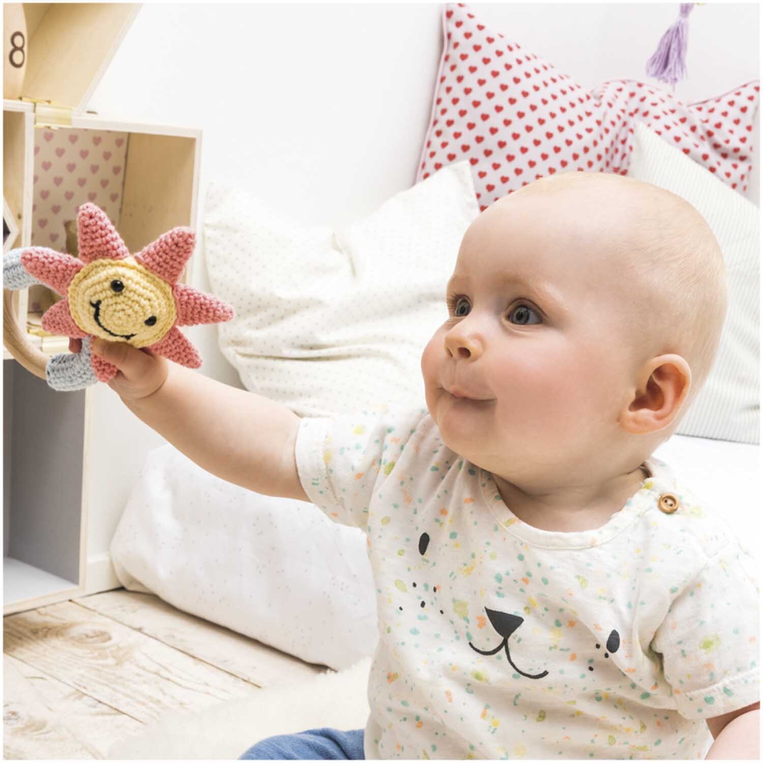 Creative Ricorumi dk: Baby spielt mit gehäkeltem Sonnenfingerpuppe in Pastelltönen, lächelnd.