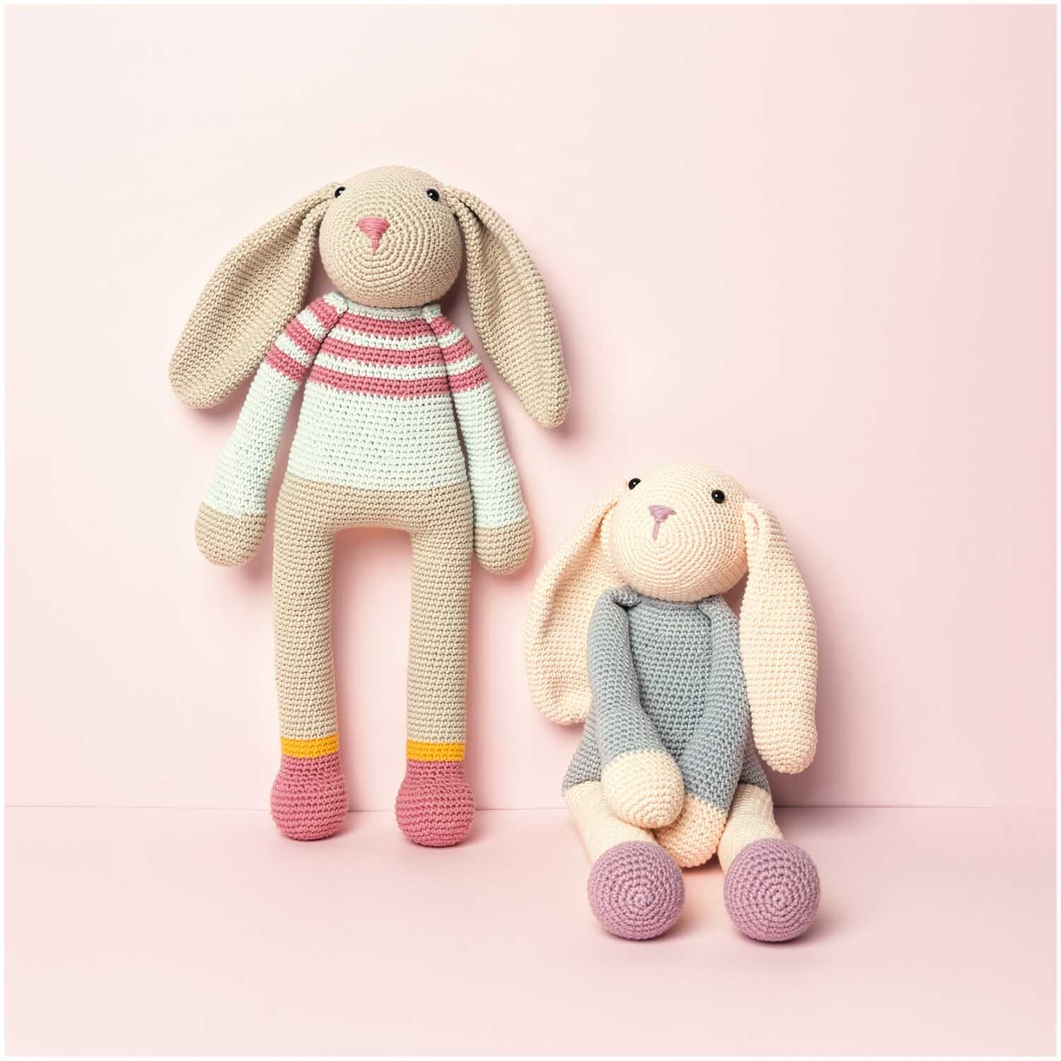 Creative Ricorumi dk: Zwei gehäkelte Hasen in Pastellfarben, einer mit gestreiftem Pullover, auf rosa Hintergrund.