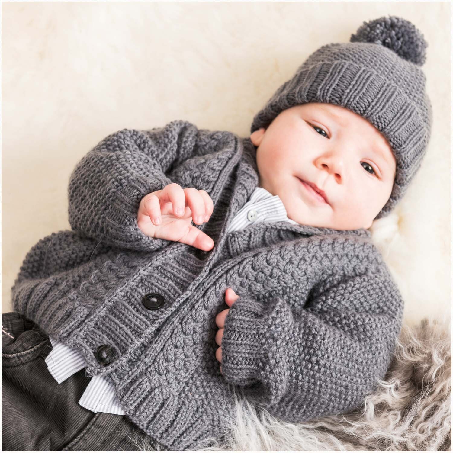 Rico Baby Heft Nr.22: Ein Baby in grauer Strickjacke und Mütze von Rico Design auf weichem Fell.