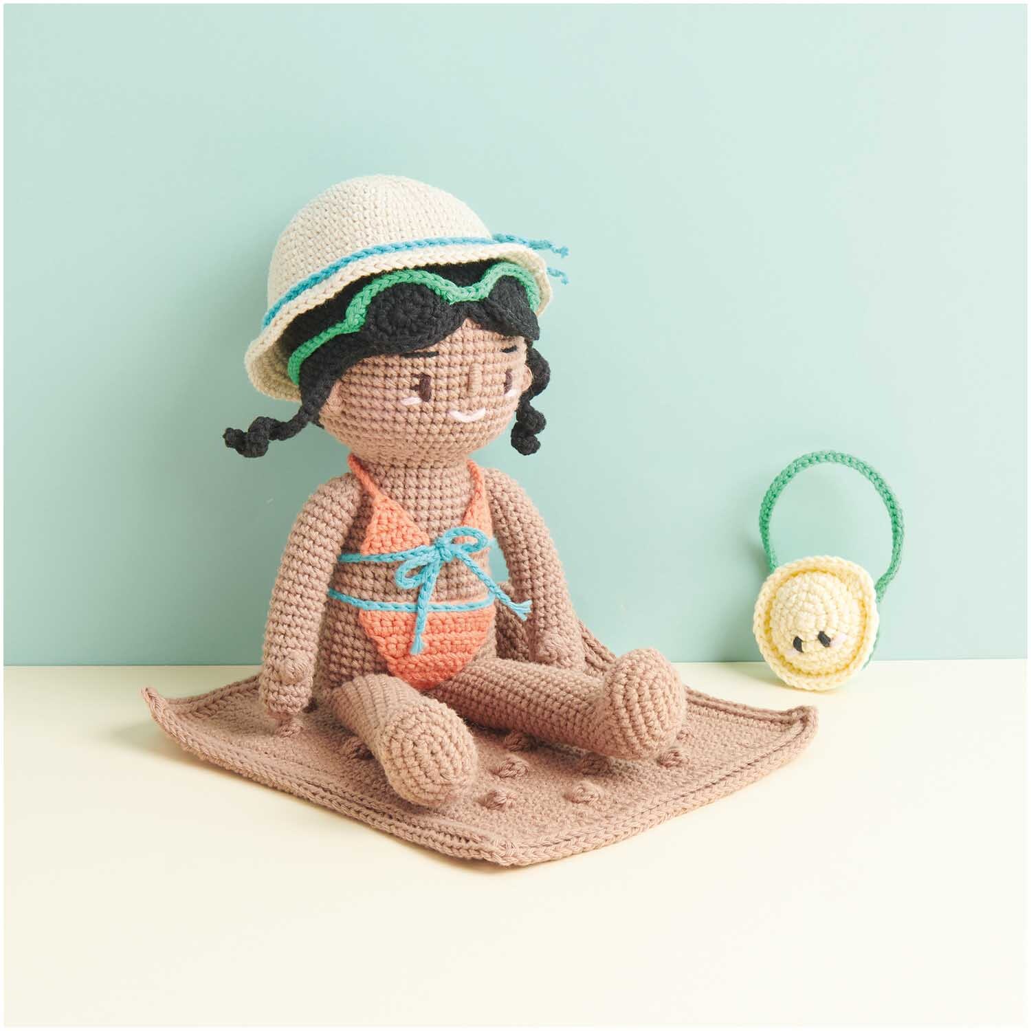 Creative Ricorumi dk: Gehäkelte Puppe mit Hut auf einer Decke, daneben ein gelbes Accessoire. Ideal für DIY-Projekte.
