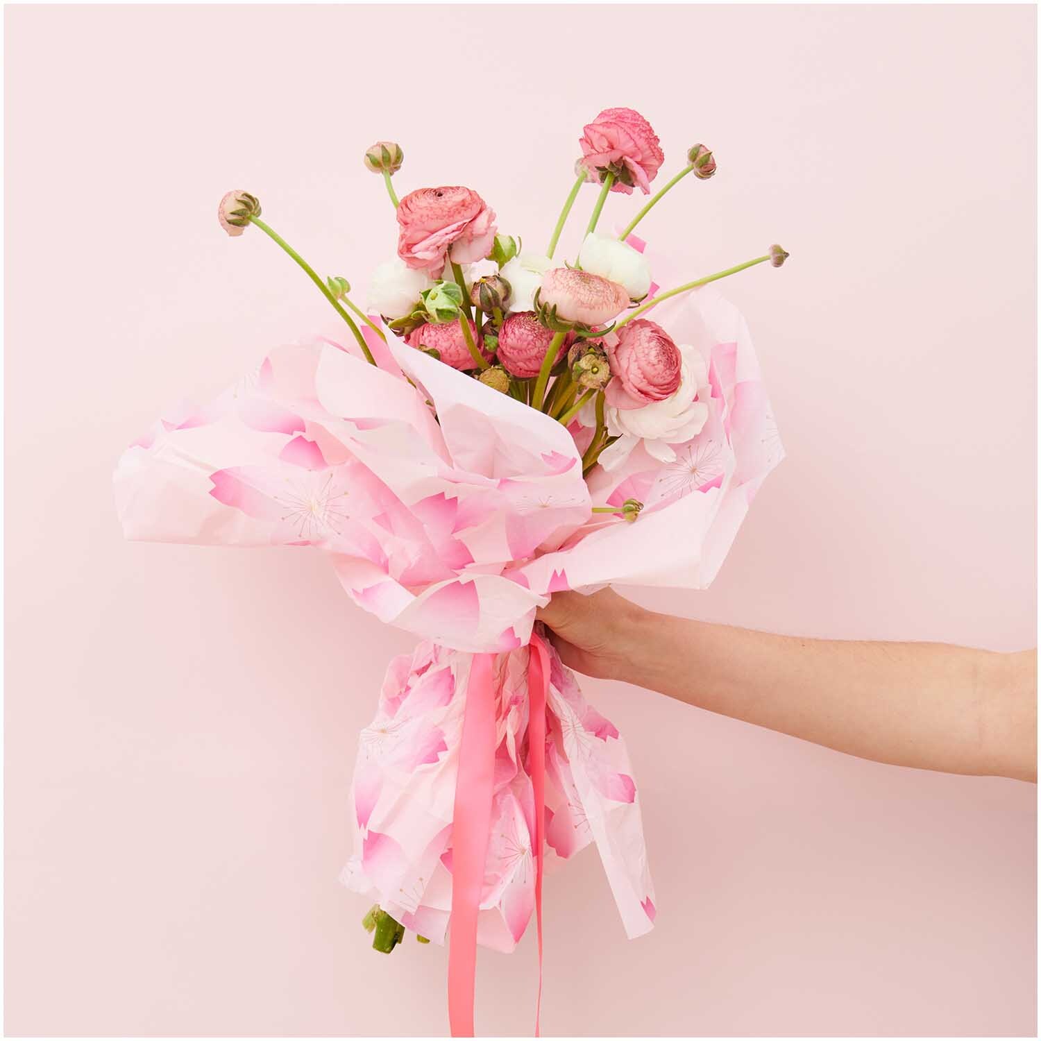 Paper Poetry Seidenpapier umhüllt einen Blumenstrauß mit rosa Blüten, gehalten von einer Hand.