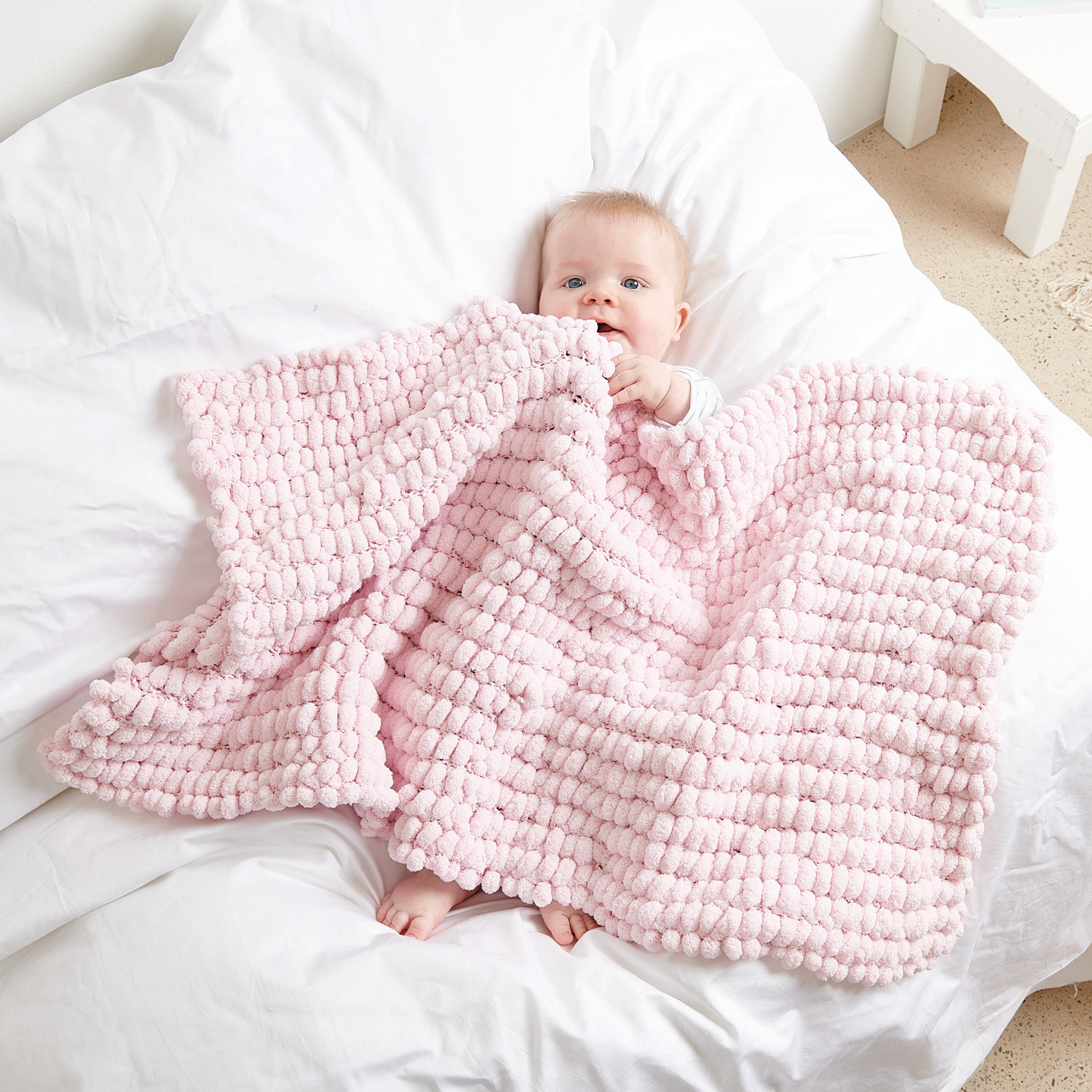 Pompon: Baby in weicher, rosa Decke von Rico Design auf weißem Bett, 200g 100m Garn.