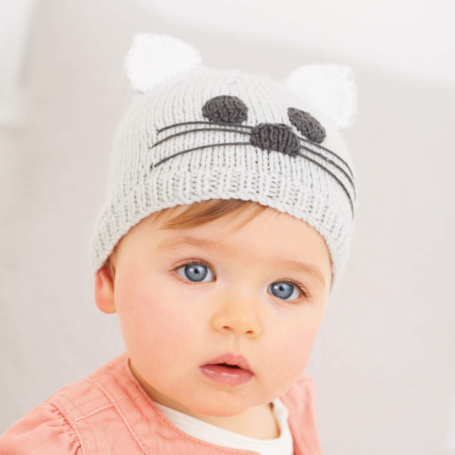 Strickanleitung Babymütze mit Katzenohren: Baby trägt graue Mütze mit weißen Ohren und Katzengesicht.