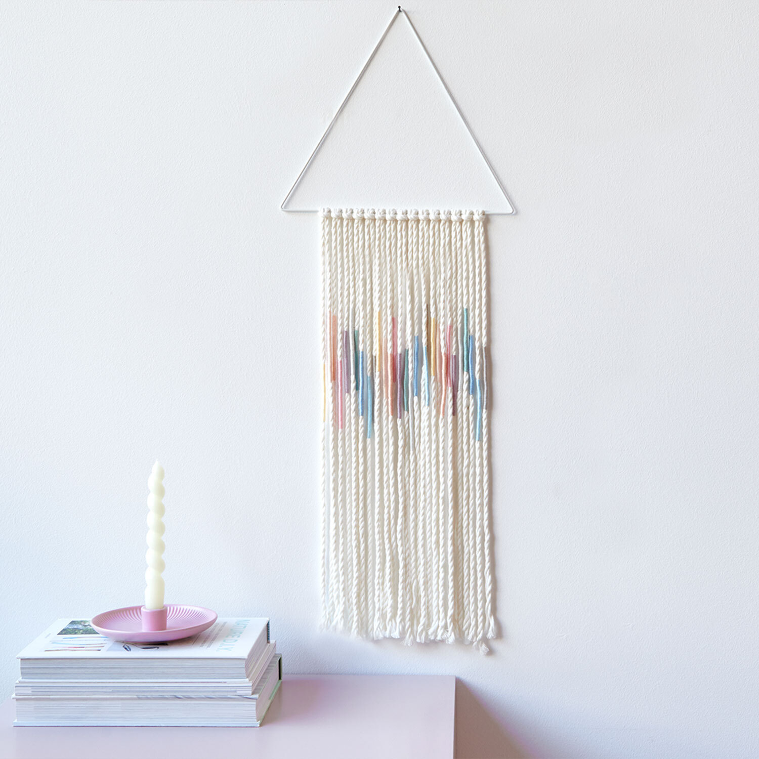 Bastelanleitung Sticktwist-Wanddeko Pastell: Weiße Wanddeko mit bunten Akzenten, hängt an dreieckigem Rahmen über Büchern und Kerze.