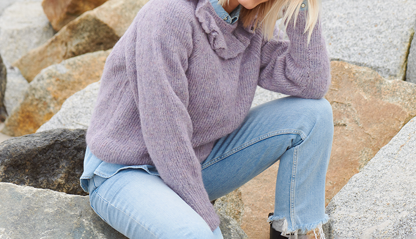 Strickanleitung Pullover mit Volant-Kragen: Frau in lila Pullover aus Alpaka-Wolle sitzt auf Felsen.