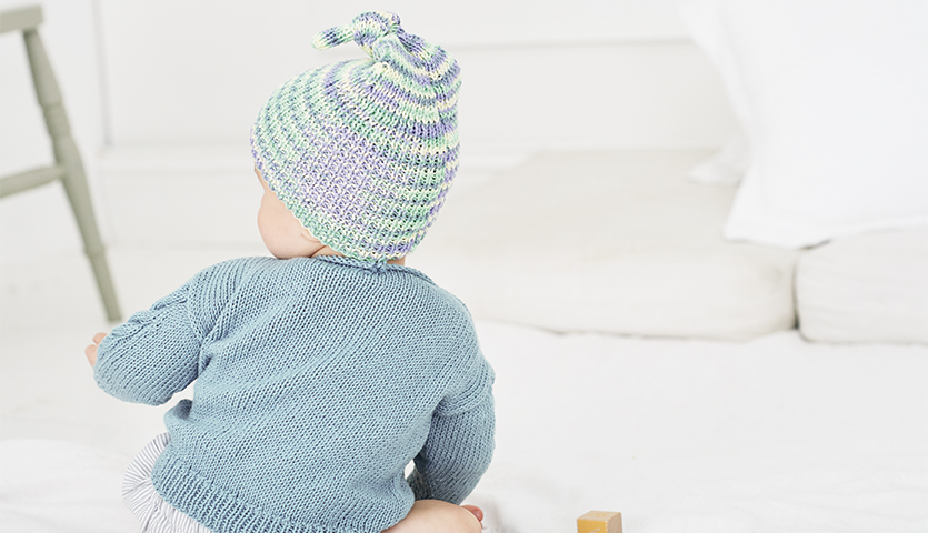 Strickanleitung Mütze/Pullover aus Baby Cotton Soft: Baby in gestrickter Mütze und Pullover in sanften Pastelltönen auf hellem Hintergrund.