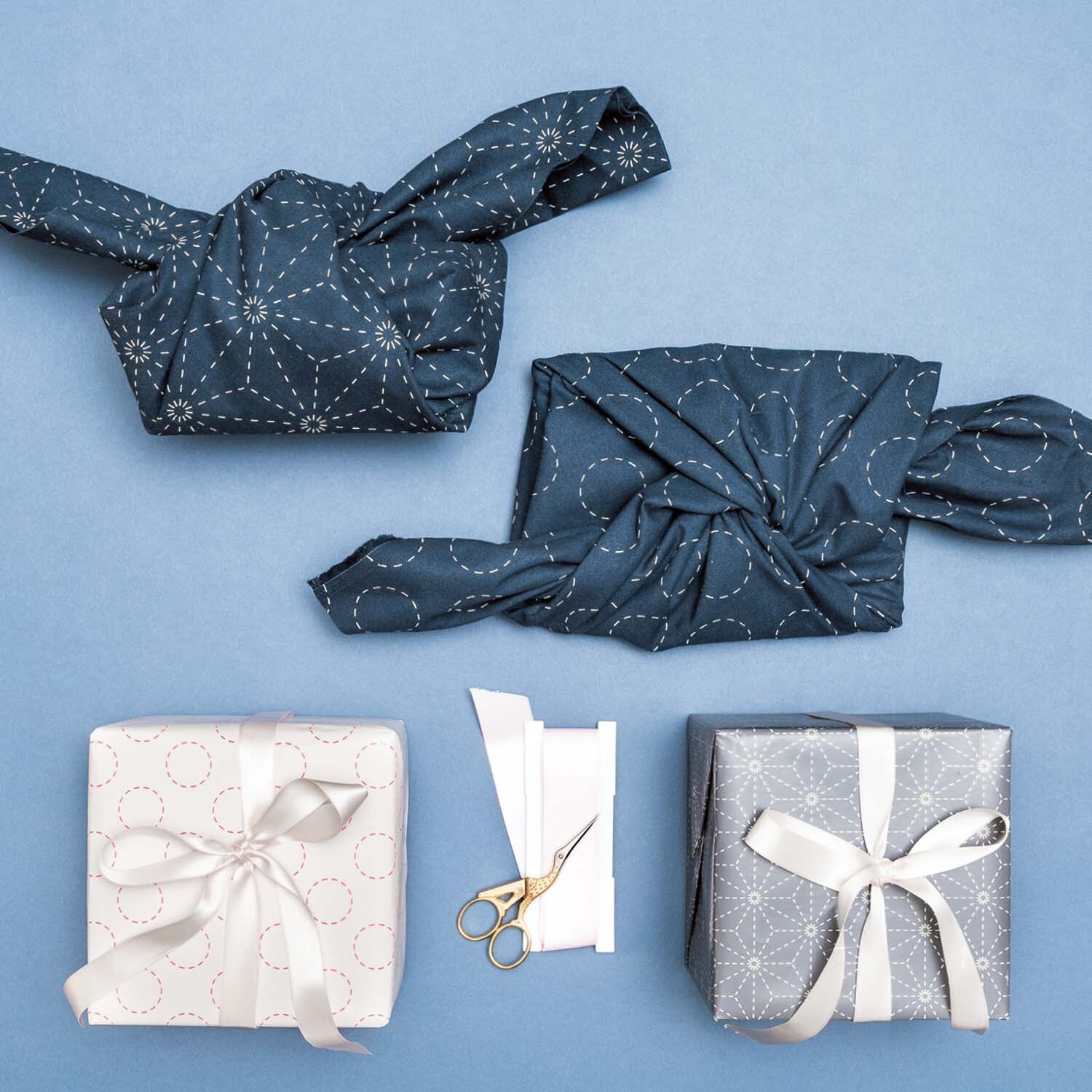 Anleitung Furoshiki: Zwei Geschenke in blauem Stoff und zwei in Papier verpackt, mit Schere.