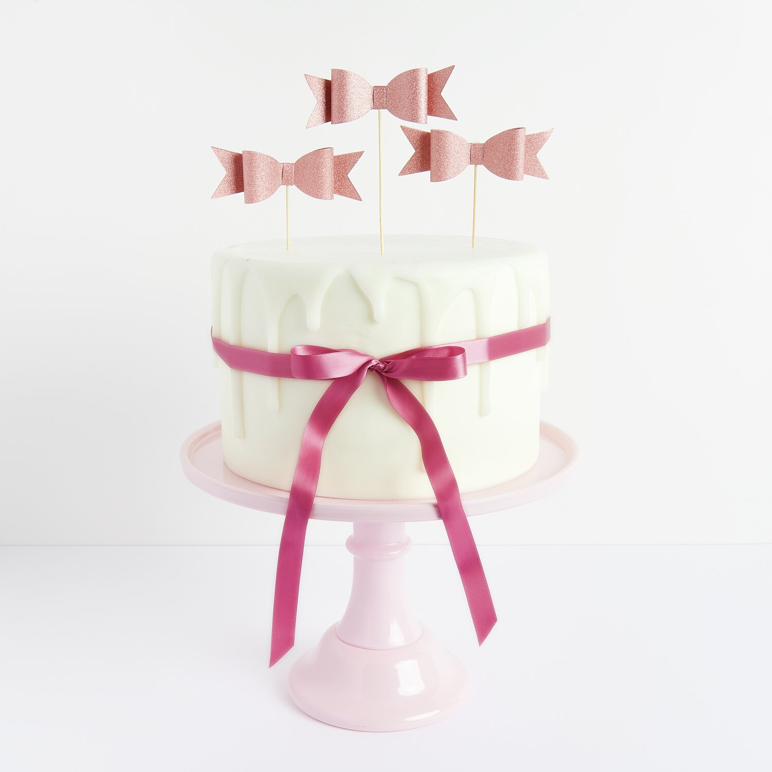 Kuchenkerzen Rosen auf einem weißen Kuchen mit roségoldener Schleife, präsentiert auf einem rosa Tortenständer.