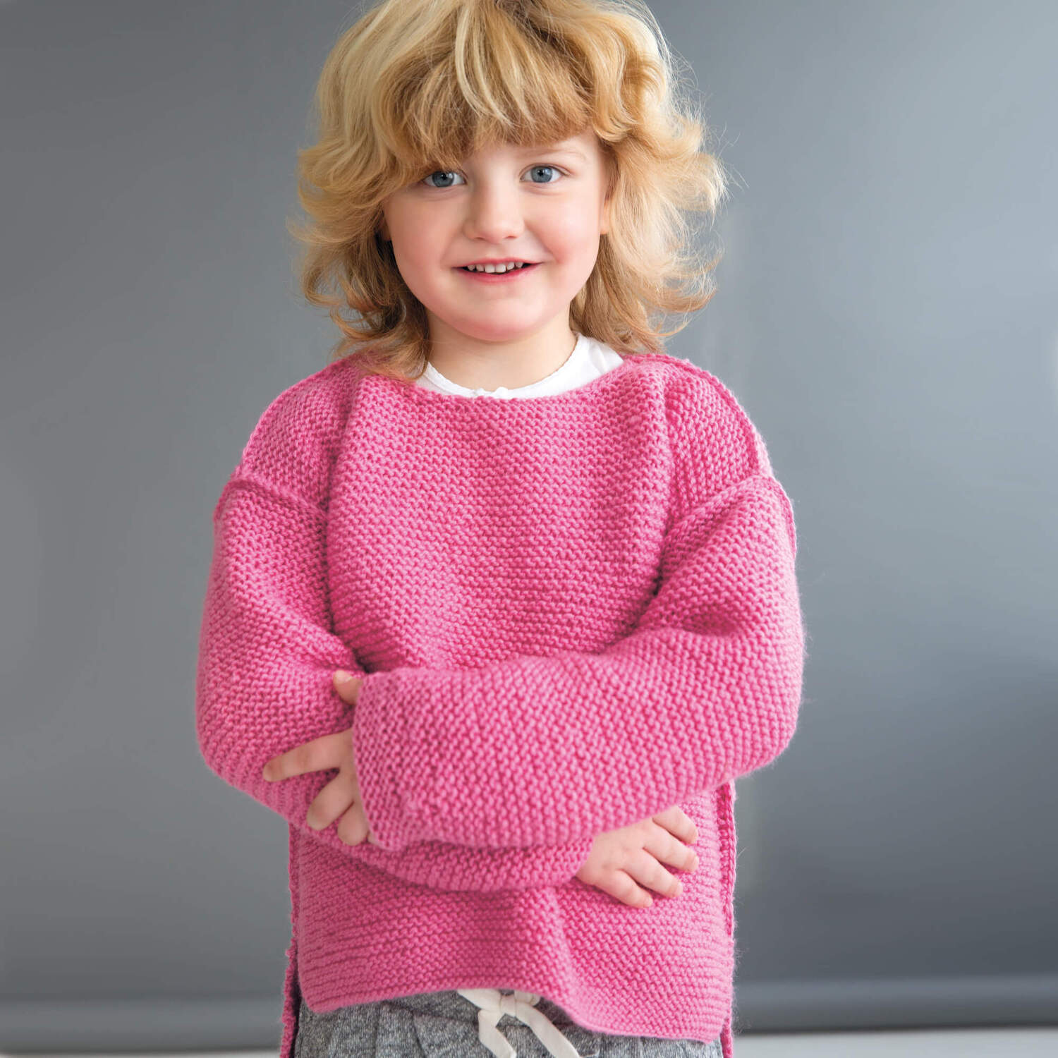 Strickanleitung Kinderpullover aus Soft Merino: Ein Kind trägt einen rosa Pullover vor grauem Hintergrund.