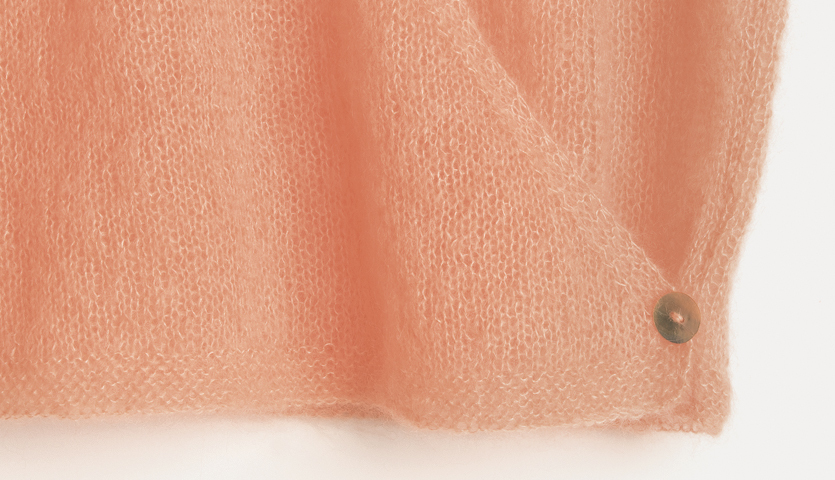 Strickanleitung Pullover in Wickeloptik: Nahaufnahme eines weichen, pfirsichfarbenen Mohair-Seiden-Stoffs mit Knopfdetail.