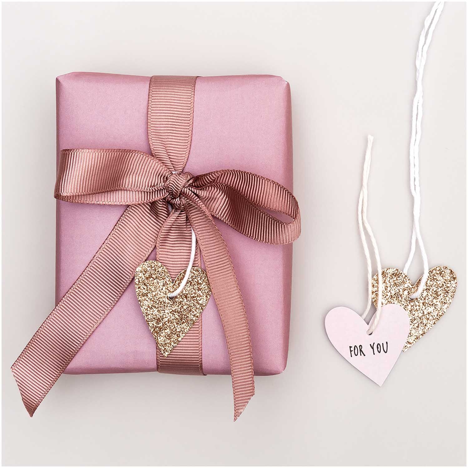 Paper Poetry Papieranhänger in Herzform mit Glitter in Roségold neben rosa Geschenk mit Schleife.