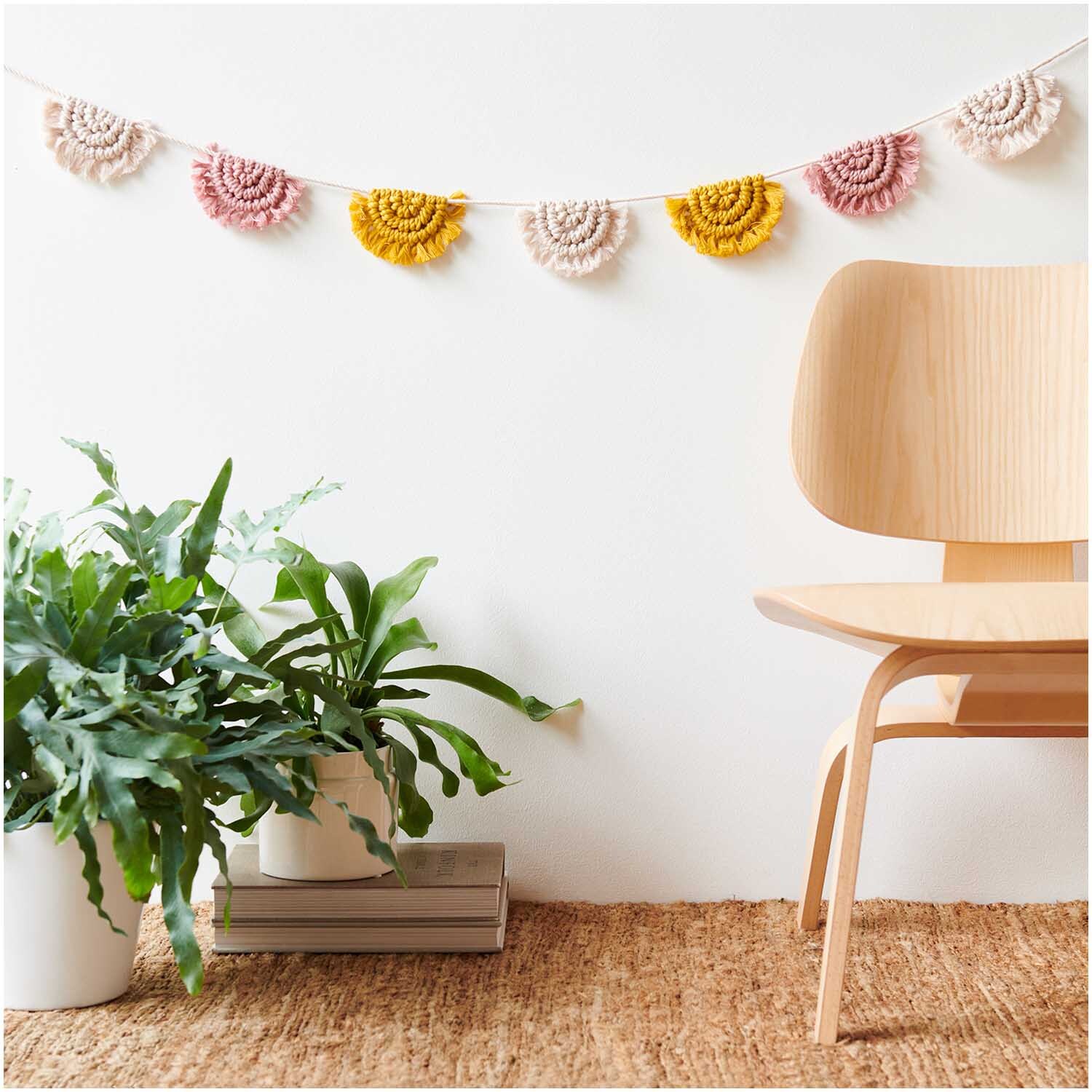 Creative Cotton Cord Makramee-Garn von Rico Design dekoriert eine Wand mit bunten Makramee-Girlanden über einem Holzstuhl und Pflanzen.