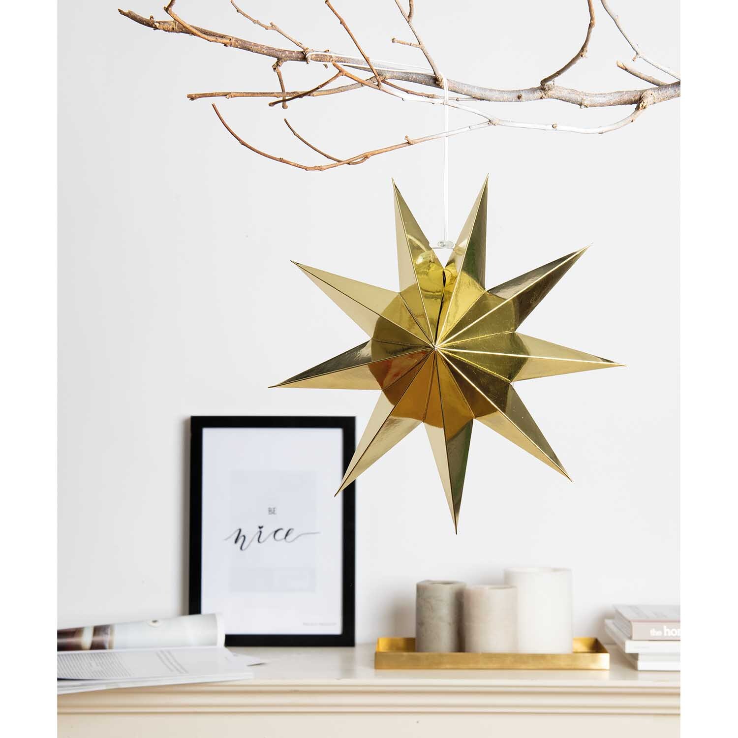 Papierstern auffaltbar 28cm, gold glänzend, hängt an einem Ast über einem Tisch mit Kerzen und einem gerahmten Bild.