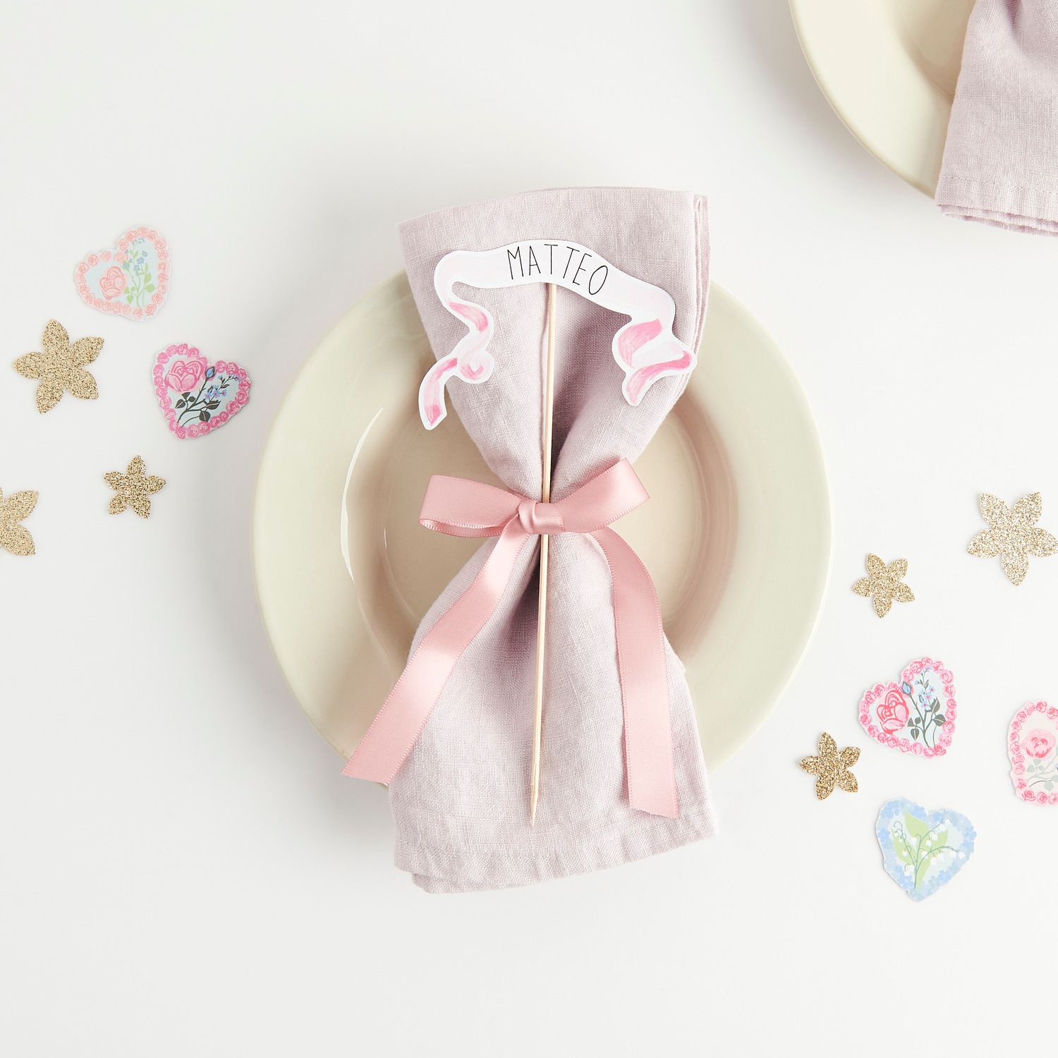 Paper Poetry Motivpapierblock La Vie En Rose: Tischdekoration mit rosa Schleife und Namensschild auf Serviette, umgeben von dekorativen Papiersternen und Herzen.
