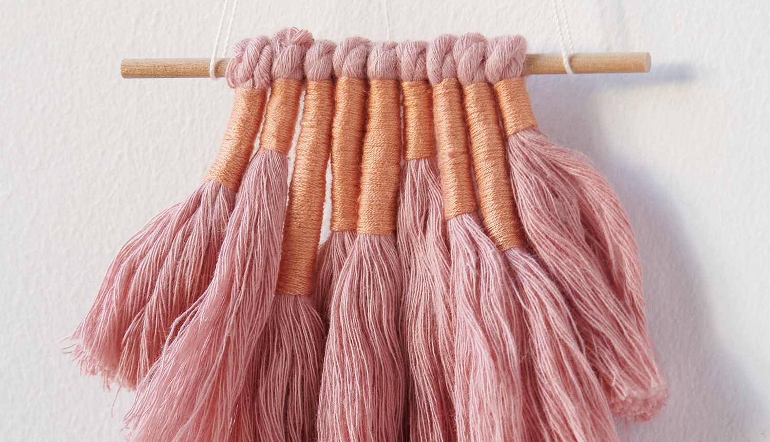 Bastelanleitung DIY Wandbehang in Erdtönen: Handgefertigter Wandbehang mit rosa Fransen an Holzstab.