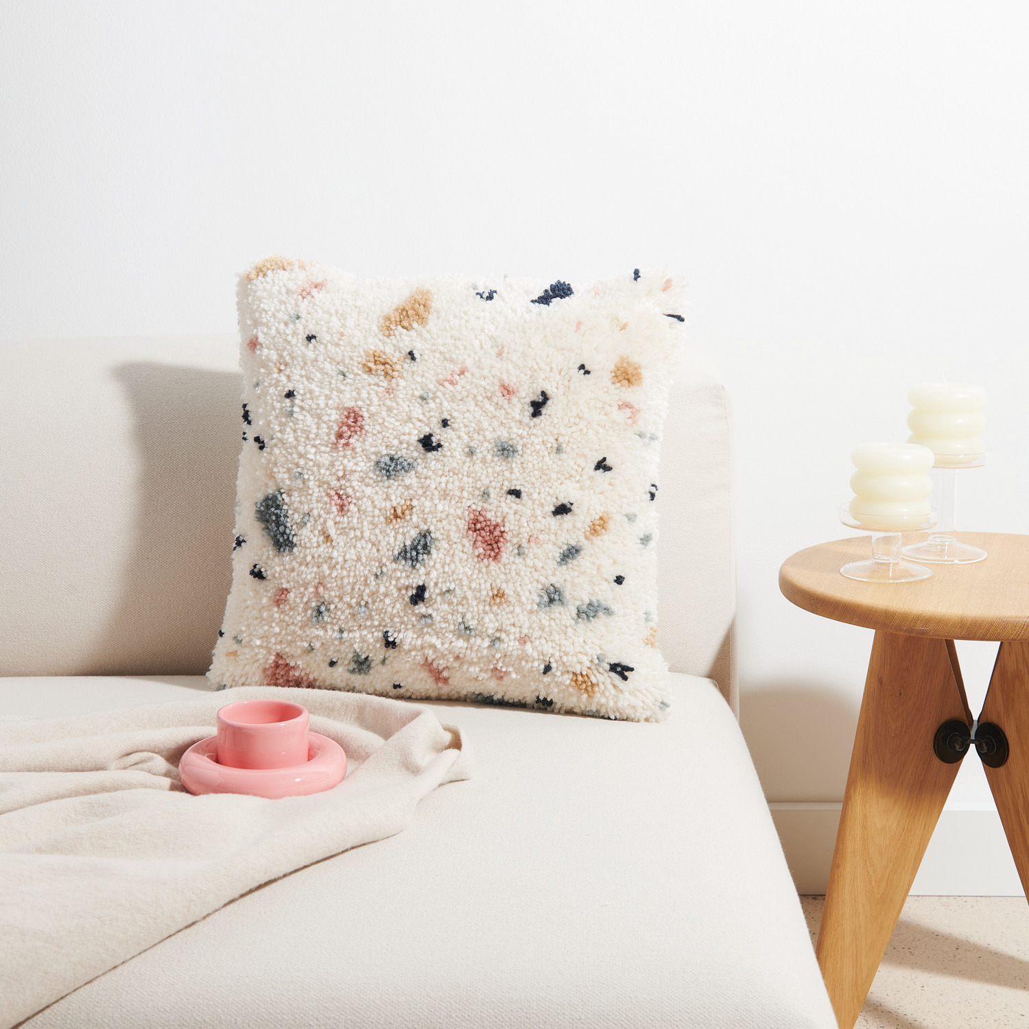Rico Hooki Hooki Knüpfset Kissen Terrazzo auf einem Sofa, mit buntem Terrazzo-Muster, 40x40cm. Daneben ein Holztisch mit Kerzen.
