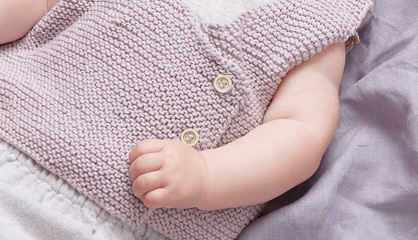Strickanleitung Weste aus Baby Cotton Soft dk: Baby trägt eine gestrickte, hellviolette Weste mit Knöpfen, auf einem lila Tuch liegend.