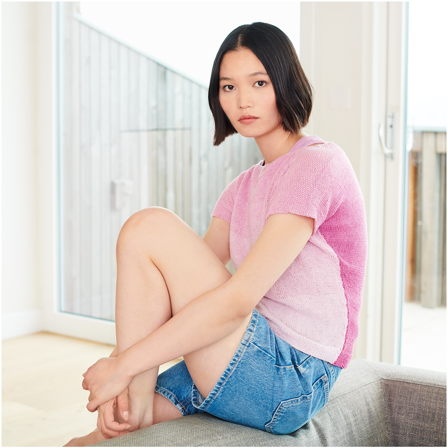 Strickset Shirt Modell 09 aus Lovewool Nr. 18: Frau in rosa Shirt und blauen Shorts sitzt auf Sofa.