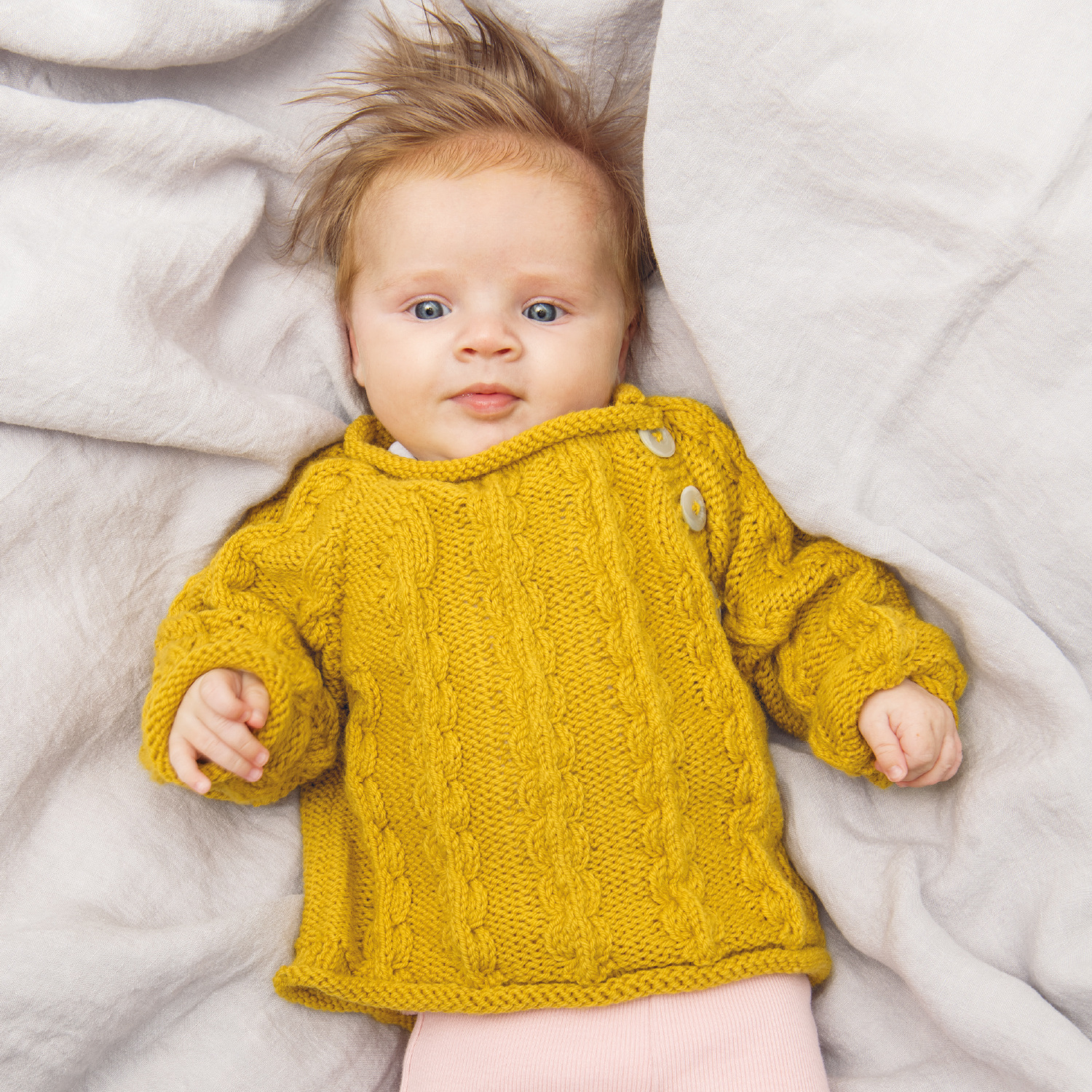 Strickanleitung Pullover aus Baby Classic DK: Ein Baby trägt einen senfgelben Strickpullover mit Zopfmuster auf einer hellen Decke.