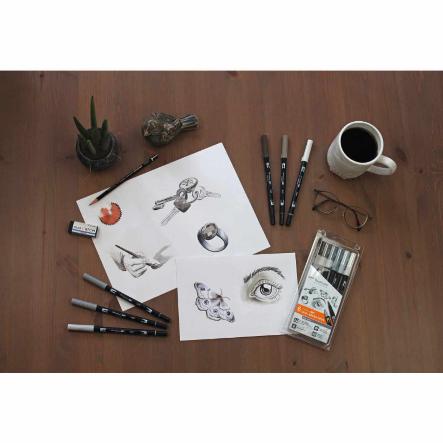 ABT Dual Brush Pen-Set Gray Colours, 6-teilig, auf einem Tisch mit Zeichnungen, Stiften und einer Kaffeetasse arrangiert.