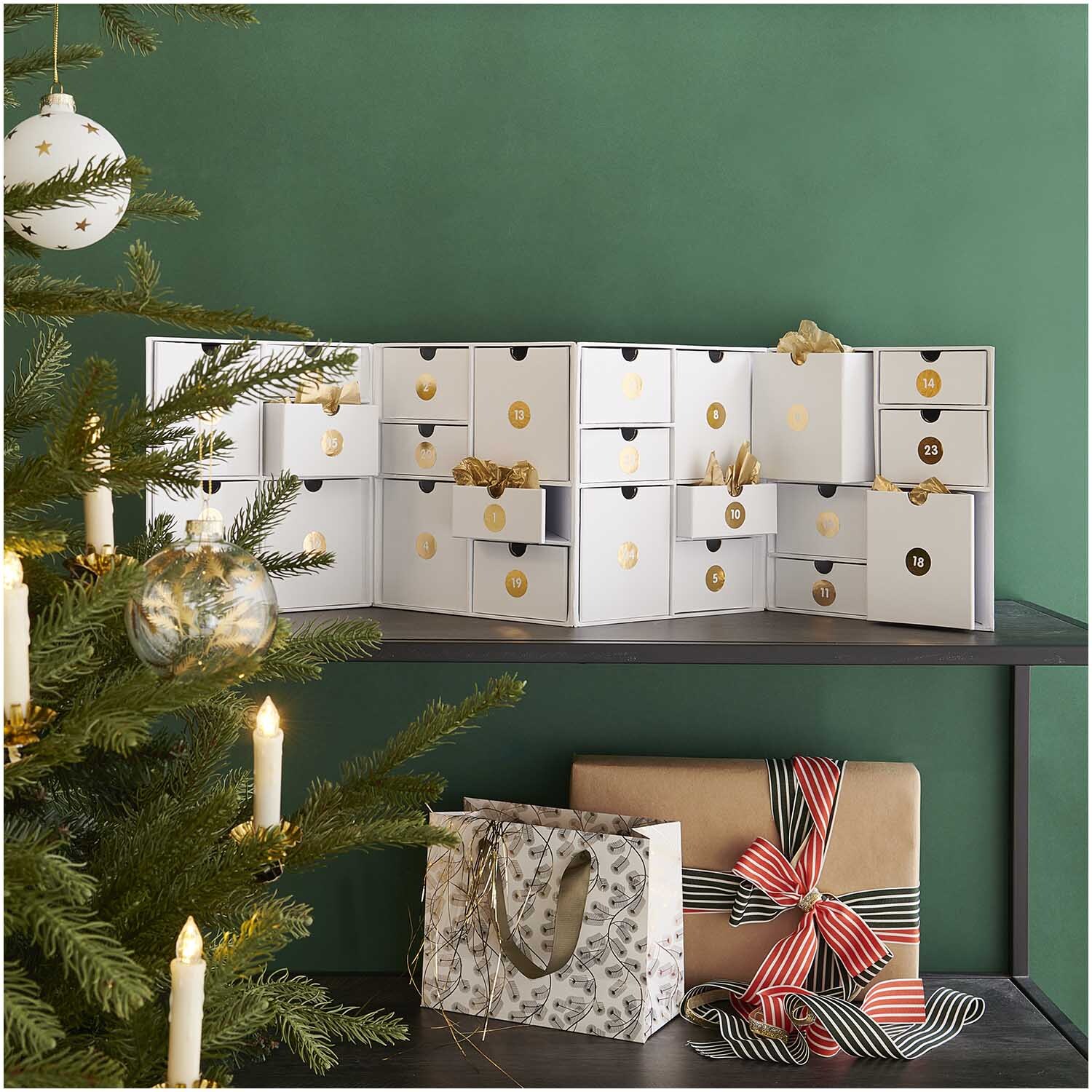 Schubladenschränkchen zum Aufklappen auf Regal, dekoriert mit goldenen Akzenten, neben Weihnachtsbaum. Ideal für festliche Aufbewahrung.